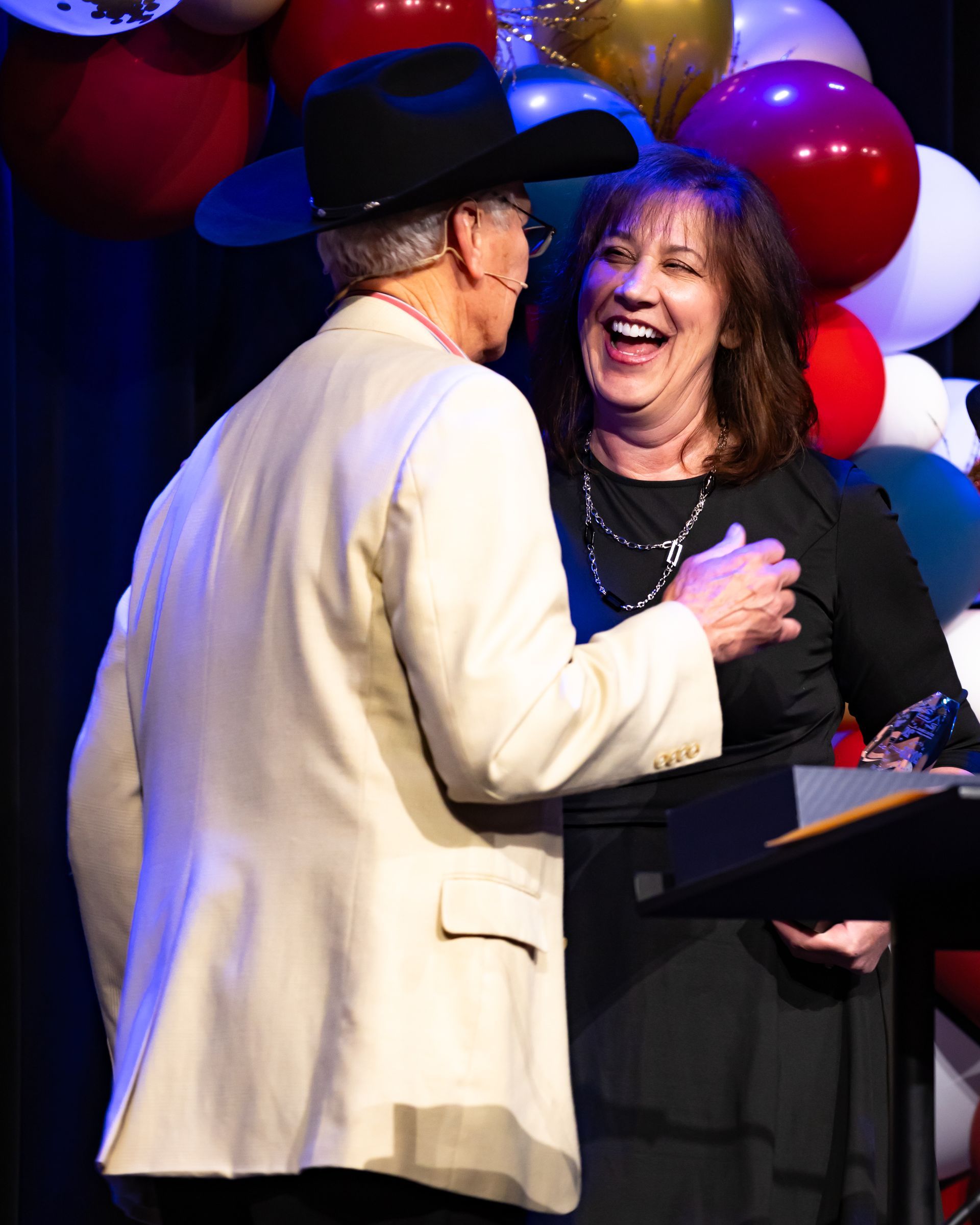 A man and a woman are laughing on a stage with balloons in the background
