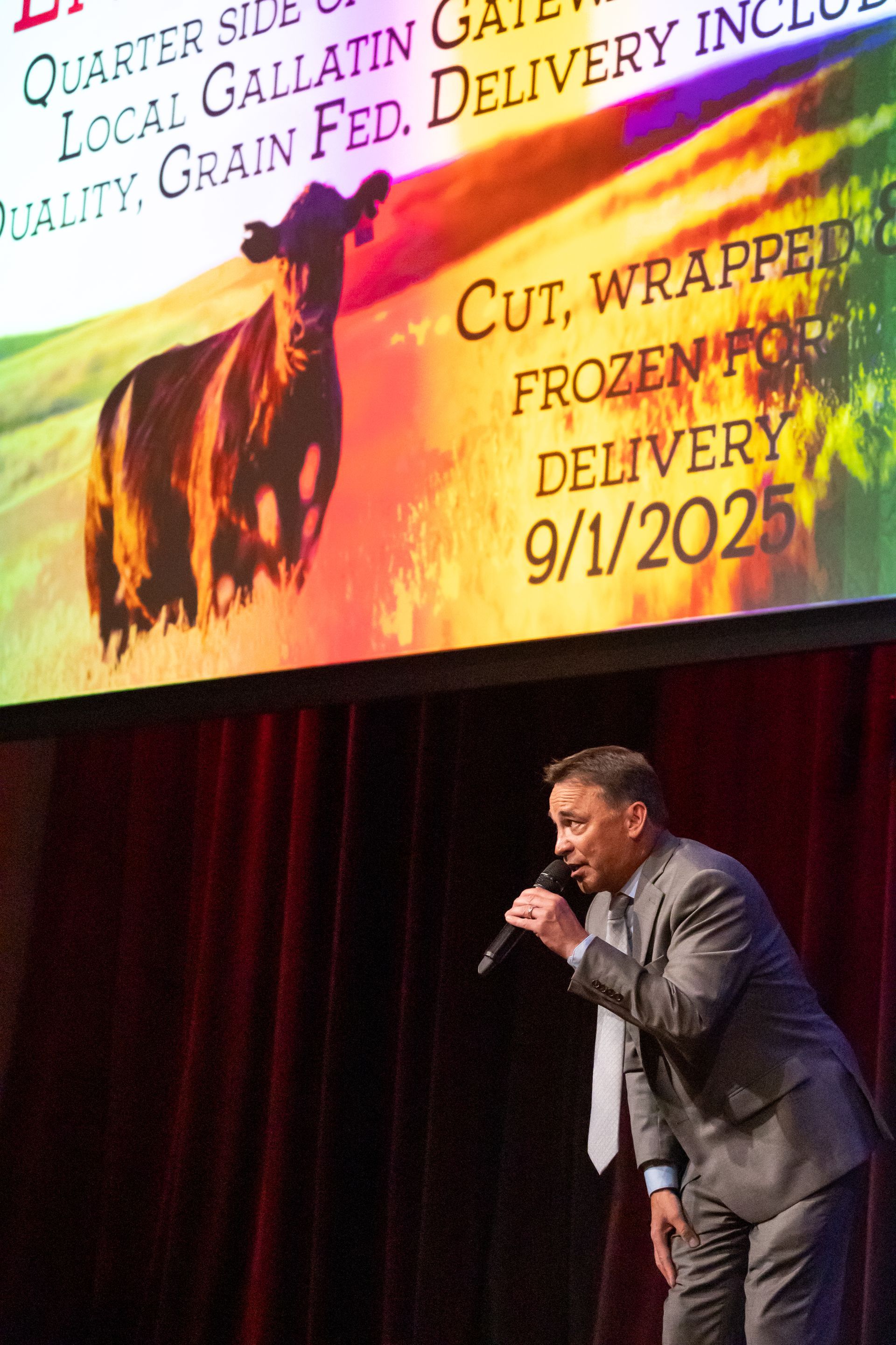 A man in a suit is singing into a microphone in front of a large screen with a cow on it.