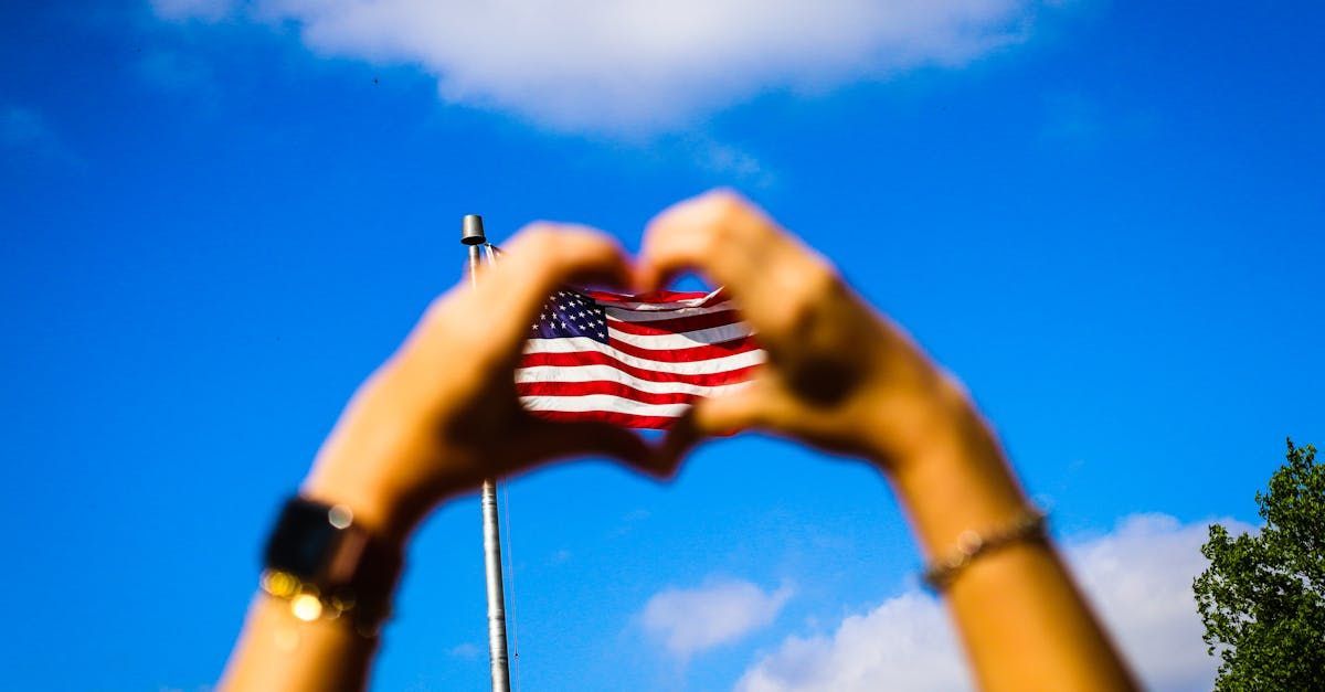 A person is making a heart shape with their hands in front of an american flag.