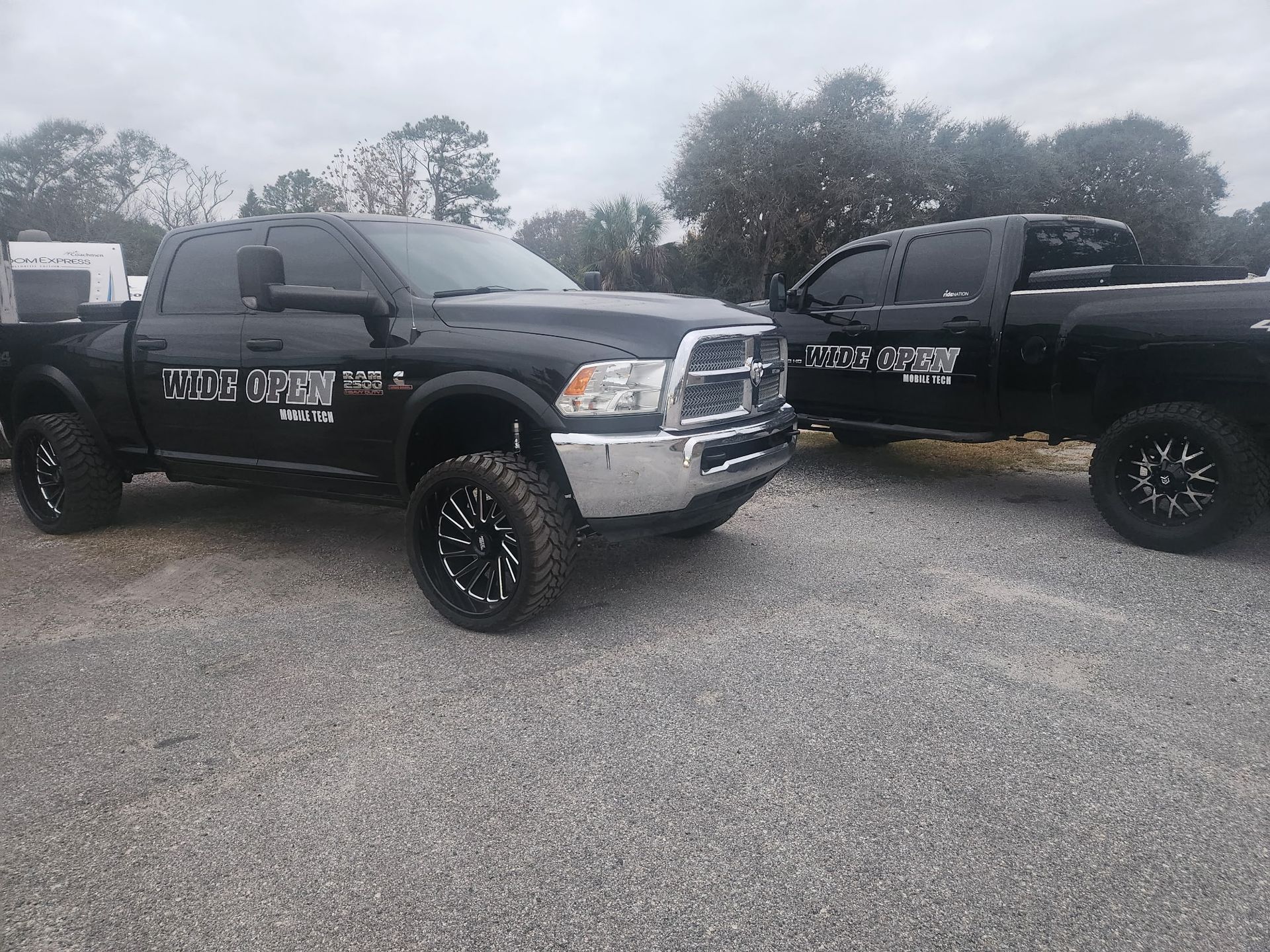Two black pickup trucks parked on gravel; both have company logos. Cloudy sky in the background.