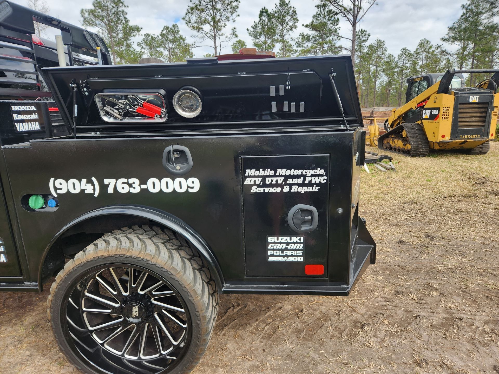 Black service truck with custom wheels, logo, phone number, and open toolbox. A yellow construction machine is in the background.