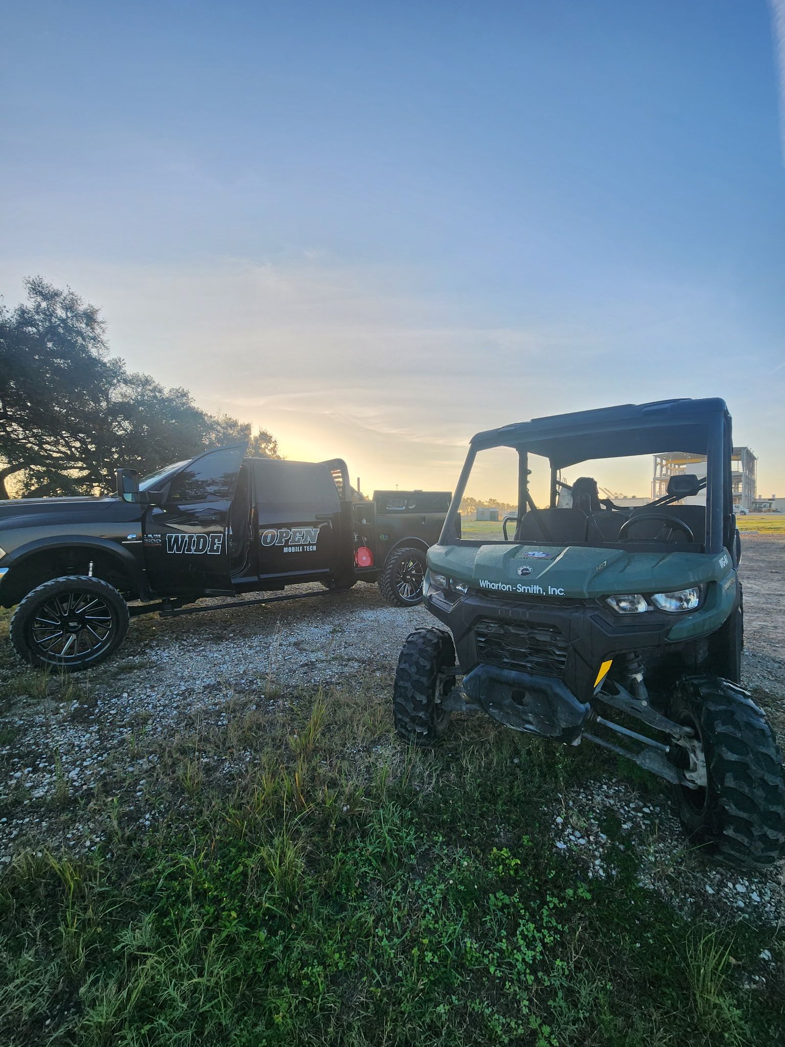 Dark truck towing a trailer and green UTV parked on a field at sunrise.