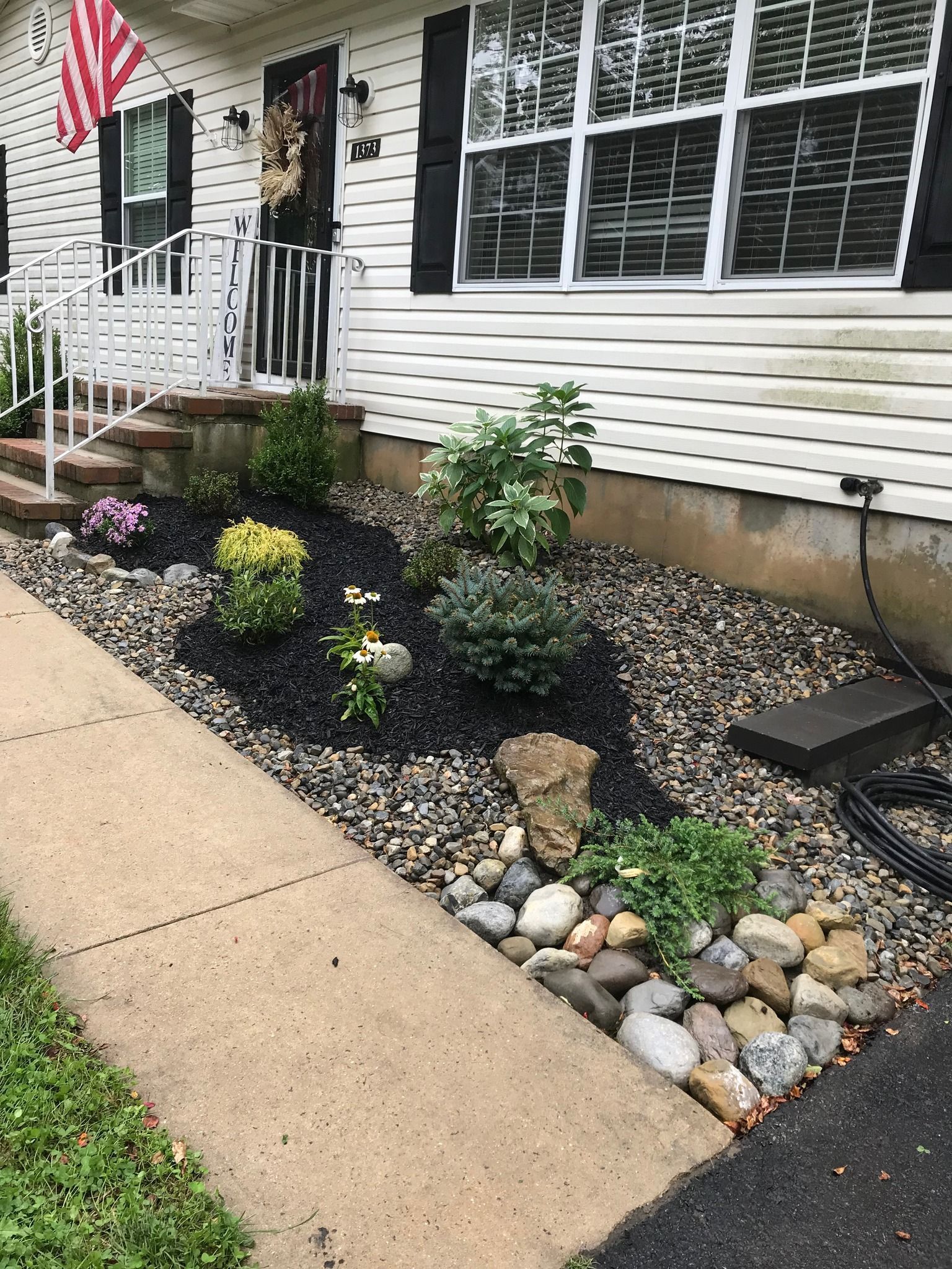 Front yard flower bed with dark mulch, river rocks, and various green plants beside a sidewalk.