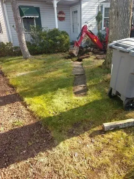 A small excavator digs in a front yard, removing a sidewalk. A house and tree are visible.