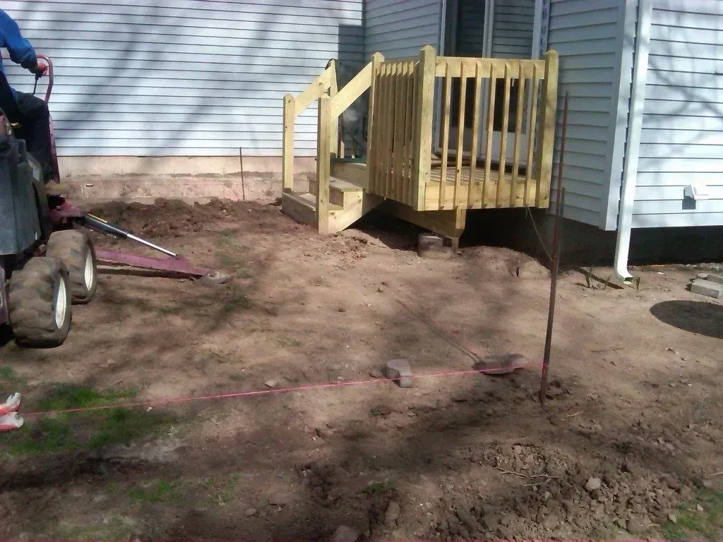 Construction site: wooden deck with stairs. A person operates a small vehicle on the dirt ground.