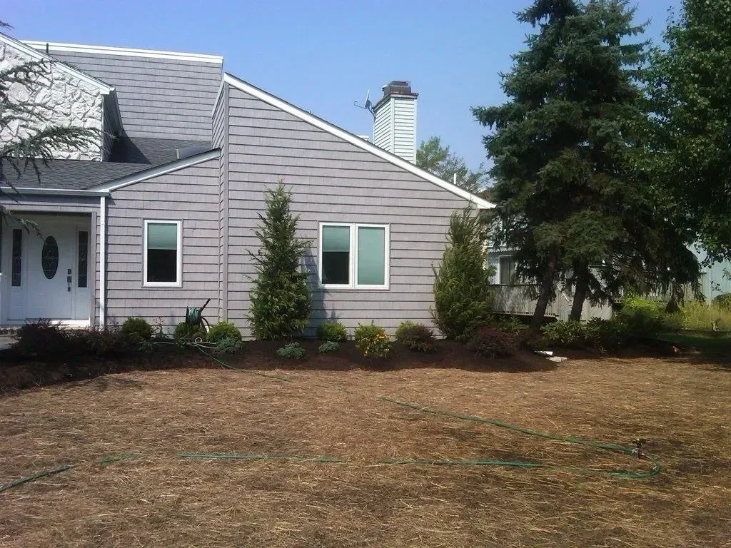 Grey house with two windows and a dark roof, surrounded by trees and a brown yard.