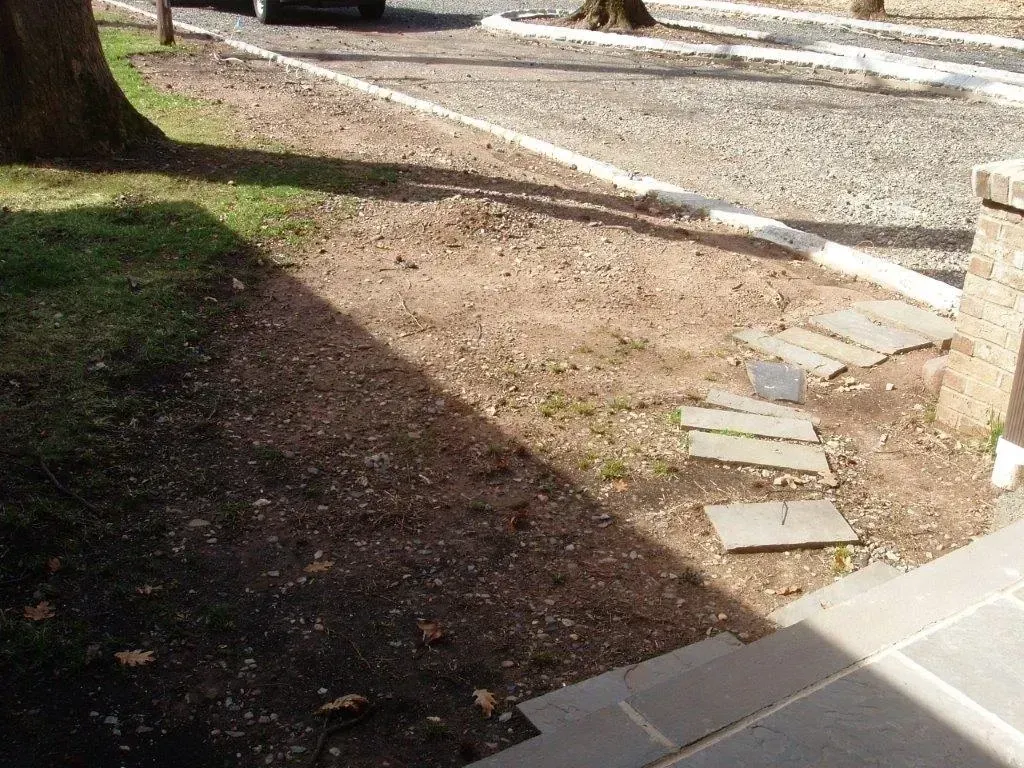 Gravel driveway next to a dirt path with stepping stones, next to a building's steps, in partial shadow.