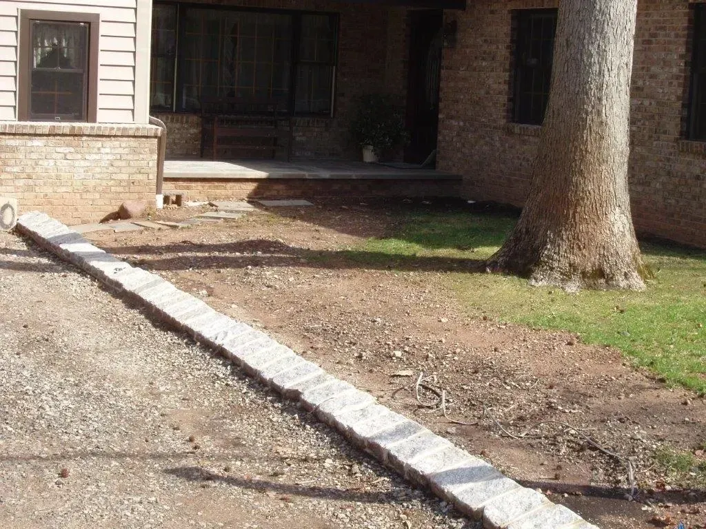 Concrete curb running along gravel driveway to a brick house with large tree.