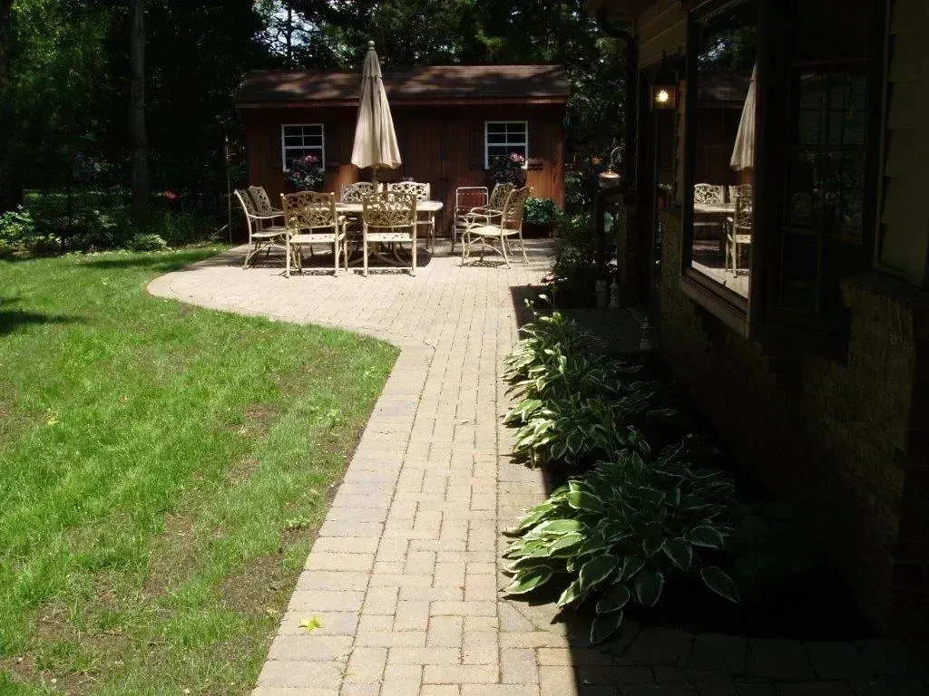 Brick pathway leading to patio with table, chairs, and umbrella. Lush green lawn and brown shed visible.