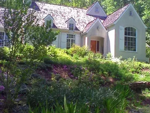 Cottage-style home with light stucco, white shutters, and a sloped roof amidst lush green landscaping.