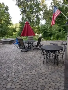 Outdoor patio with stone floor, tables, chairs, red umbrella, American flag. Trees in the background.