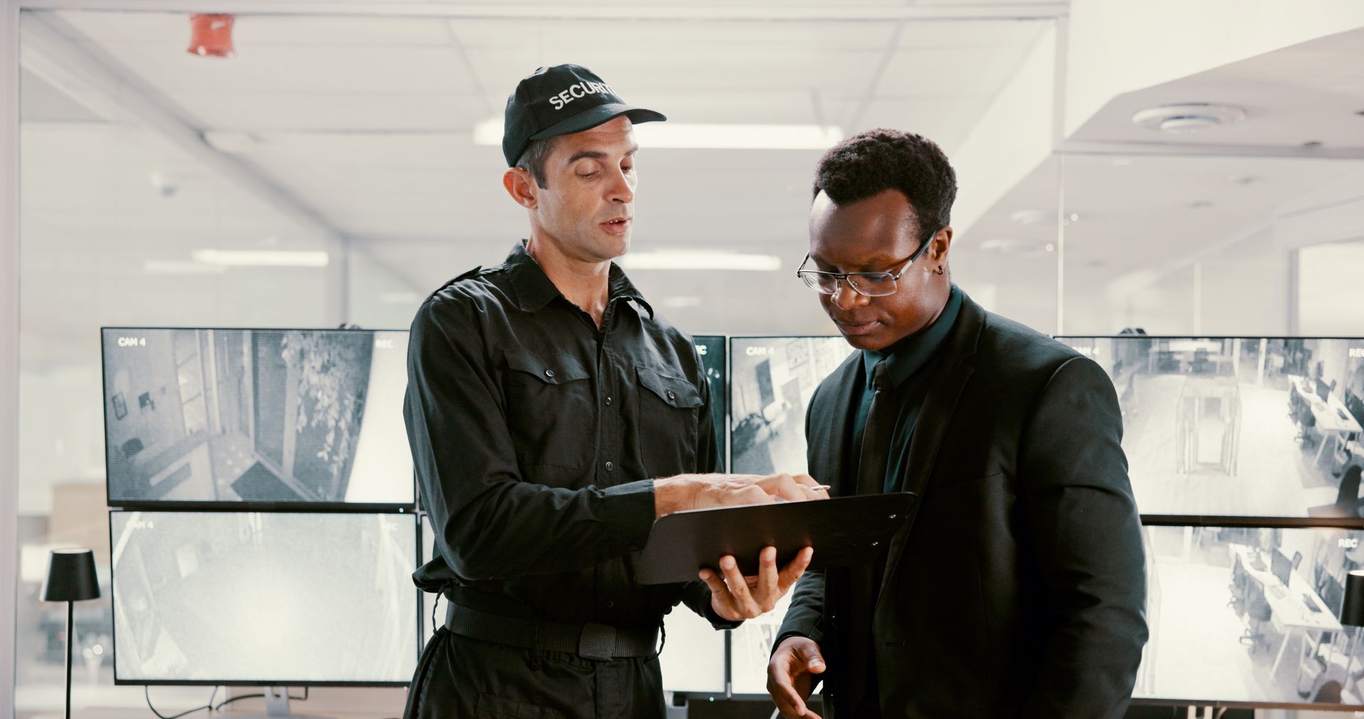 A security guard and team member are watching surveillance in a control room.