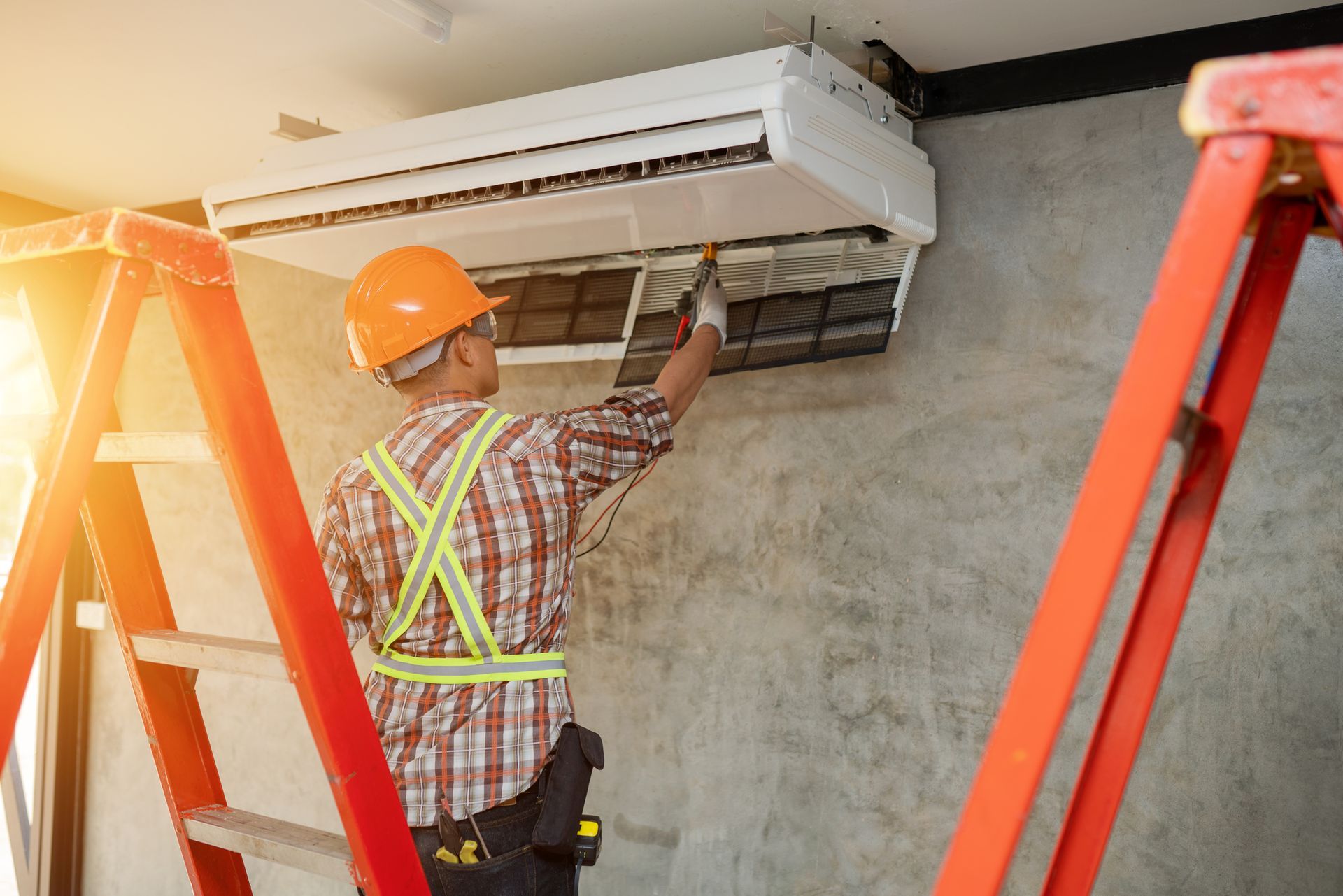 HVAC repair contractor checks on an AC unit inside a room, with orange stepladders behind him.