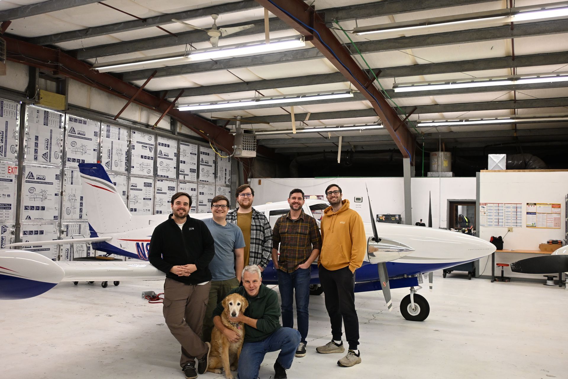 Group of people and a dog posing in front of an airplane inside a hangar.