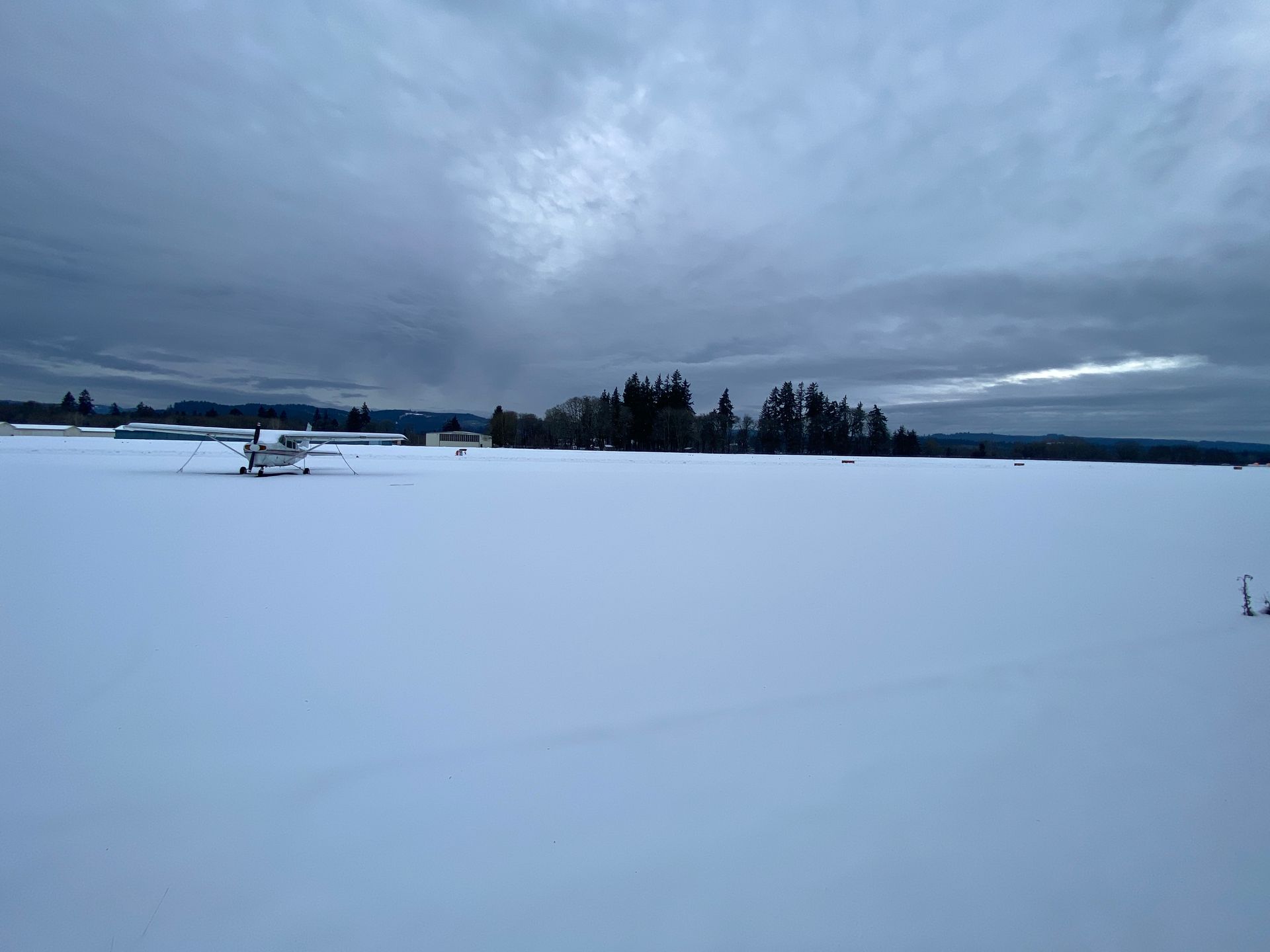 Snow-covered field under a cloudy sky. Trees and buildings are visible in the distance.