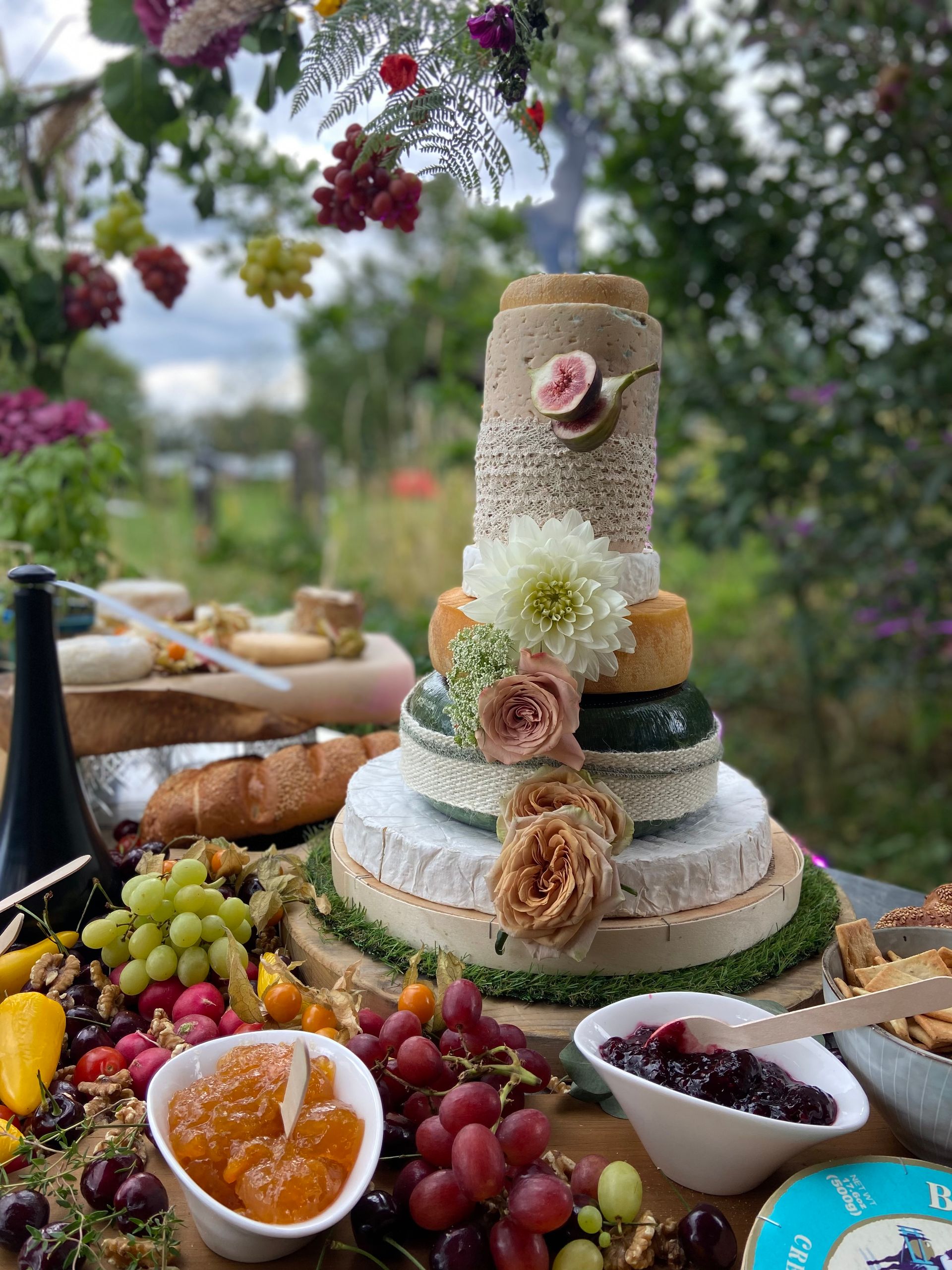 Een prachtige toren van verschillende kazen, gestapeld als een bruidstaart, versierd met verse vijgen, rozen, en een witte chrysant. De kaasopstelling is omgeven door een overvloed aan verse druiven, vijgen, stokbrood en schaaltjes met jam. De achtergrond toont een weelderige tuin met bomen en hangende druiven, wat het geheel een rustieke en romantische sfeer geeft