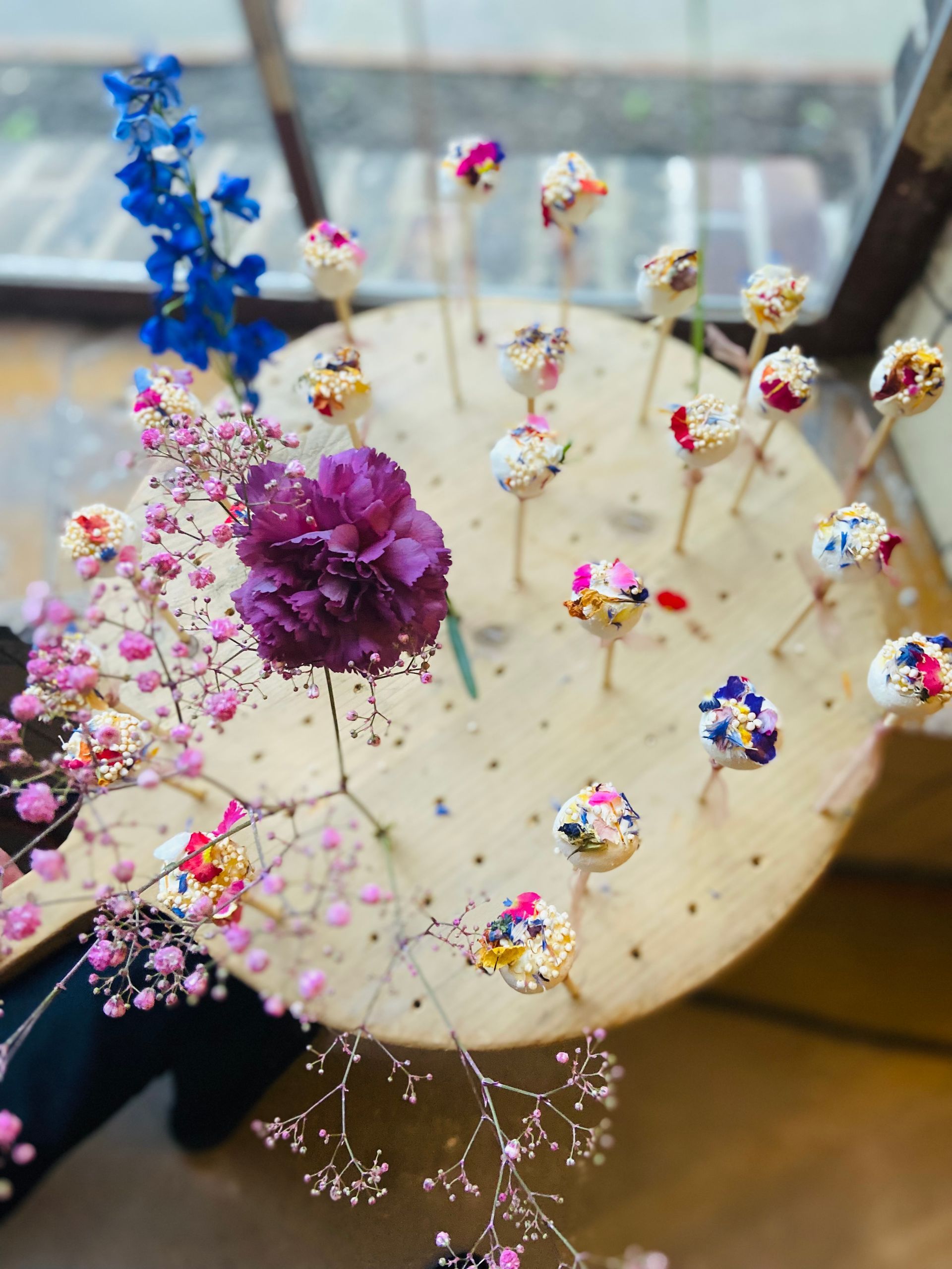 Een houten standaard met kleine gaatjes waarin kleurrijke lollie's zijn gepresenteerd. De lollie's zijn versierd met eetbare bloemen en gouden accenten, wat een luxueuze uitstraling geeft.