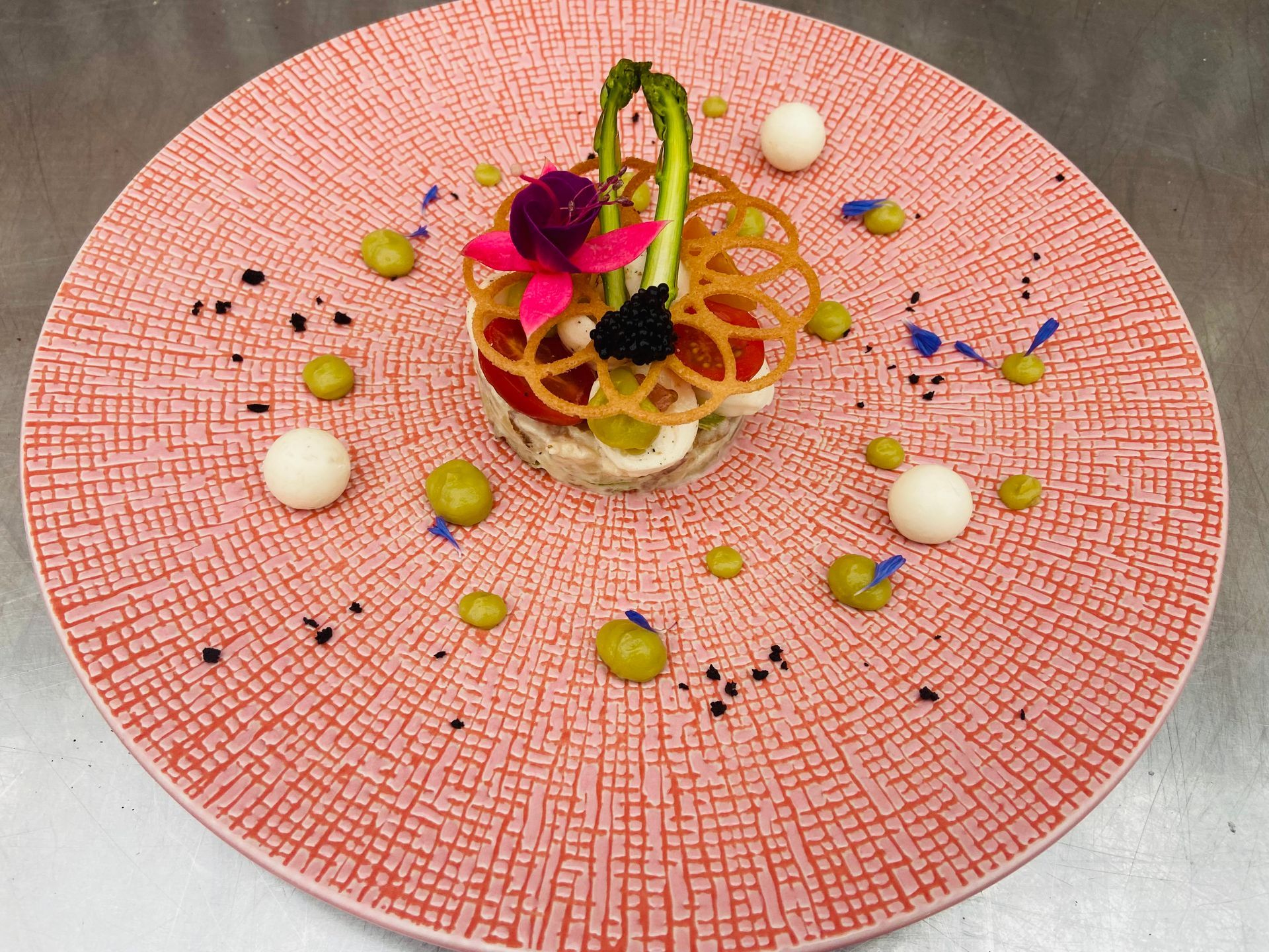 Een elegant en artistiek opgemaakt veggie gerecht geserveerd op een roze, gestructureerd bord. Het gerecht bevat een cilindervormige opbouw van ingrediënten, waaronder aspergepunten, eetbare bloemen, zwarte kaviaar, dunne krokante ringen en diverse kleurrijke elementen. Rondom het gerecht zijn kleine dotjes groene crème, witte bolletjes en blauwe bloemblaadjes geplaatst, met een subtiele toevoeging van zwarte korreltjes die het geheel verfijnen.