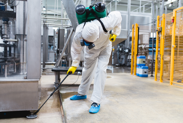Worker in white protective suit cleaning industrial equipment with a pressure washer in a factory.