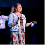 Speaker on stage holding a microphone and pointing, wearing a floral dress and denim jacket under stage lighting