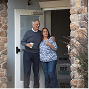Two people standing in a doorway, smiling, with a potted plant on the porch