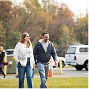Two people walking on a grassy path near parked cars in an autumn park