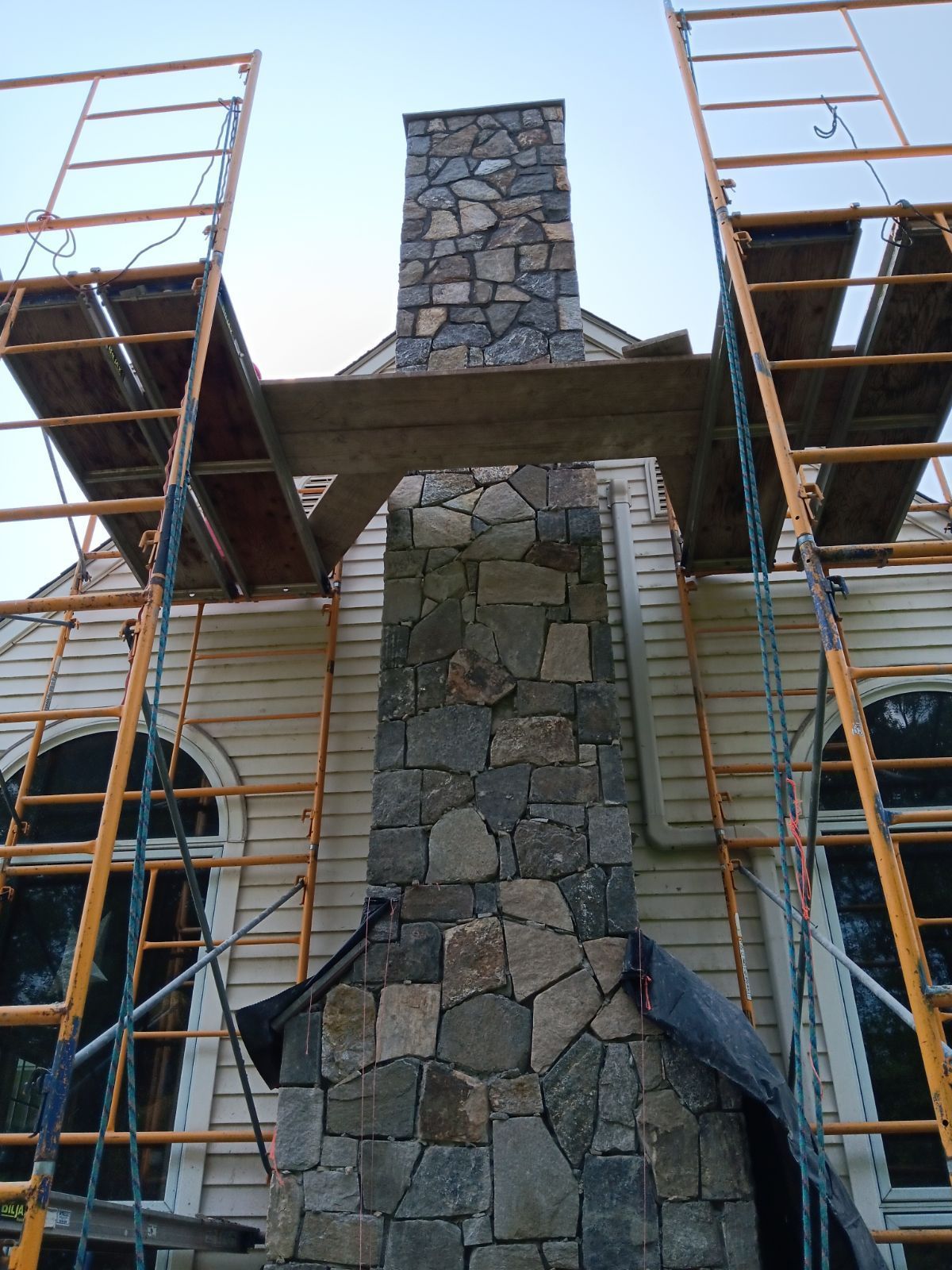 Chimney under construction, with stone facade, scaffolding, and a house in the background.