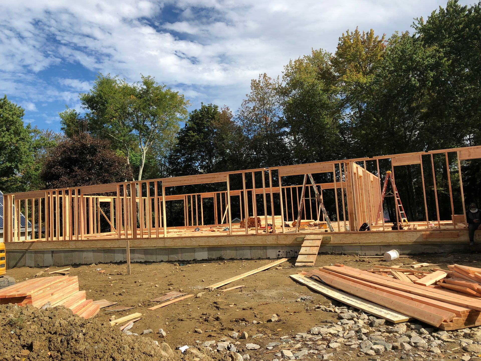Framing of a wood-framed house under construction on a concrete foundation; lumber and trees in background.