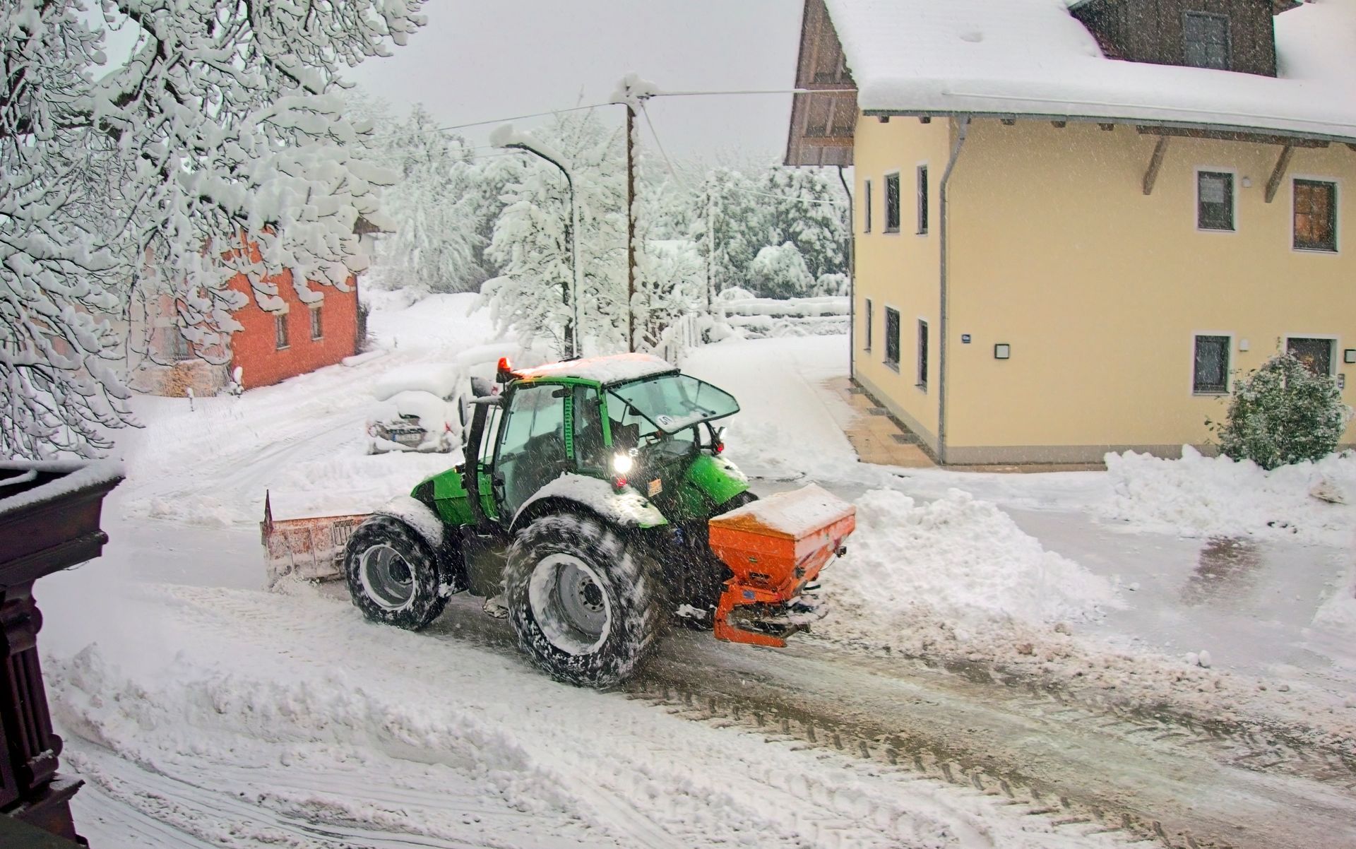 Tractor plowing snow-covered street near a yellow house and red building in a snowy, winter scene.