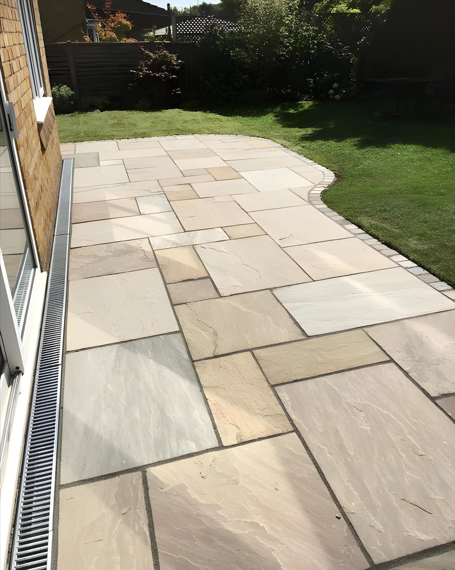Patio made of light tan stone, bordering green lawn, beside a brick wall.