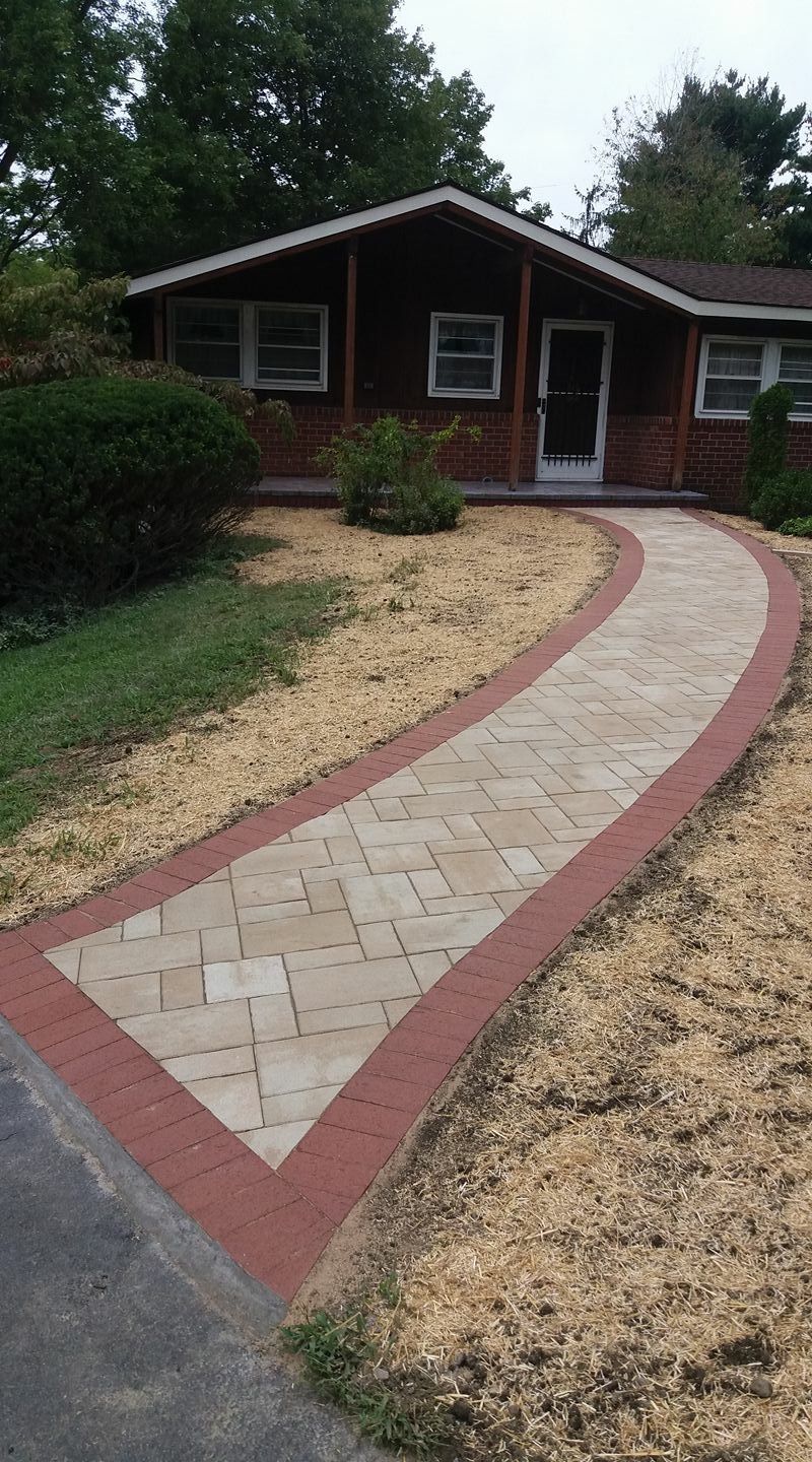 Brick walkway with red border leading to a brown house with a porch, on a sunny day.