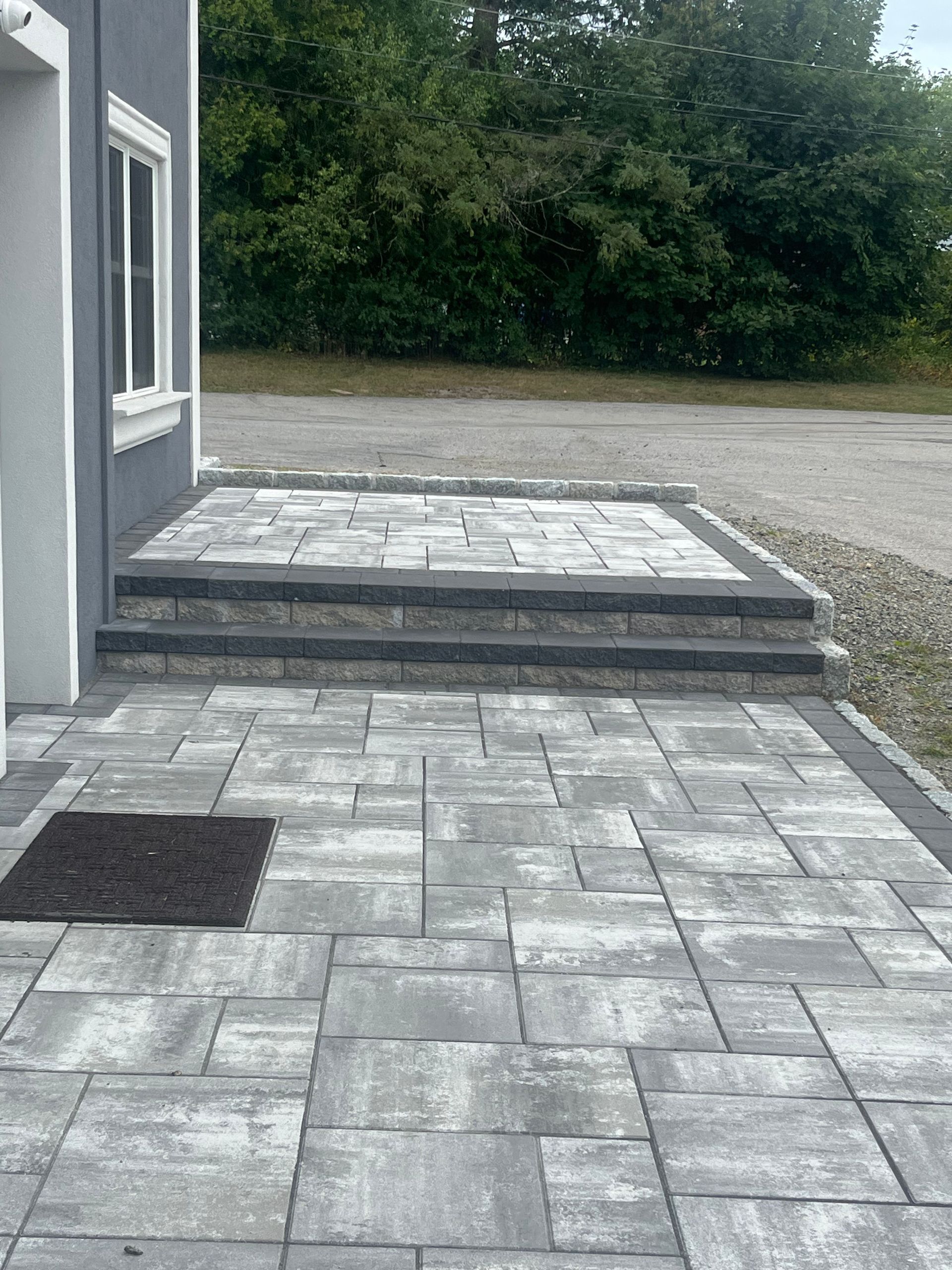 Gray paver patio with steps leading to a doorway. A welcome mat sits on the patio.