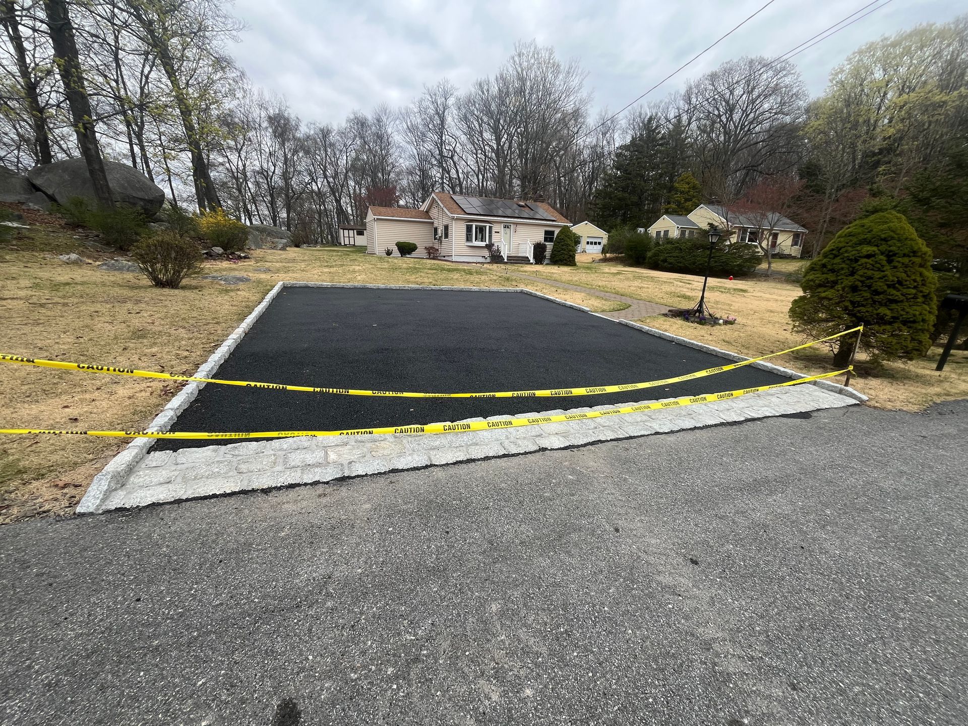 Black asphalt parking area bordered by gray stone, with yellow caution tape, in front of a house.