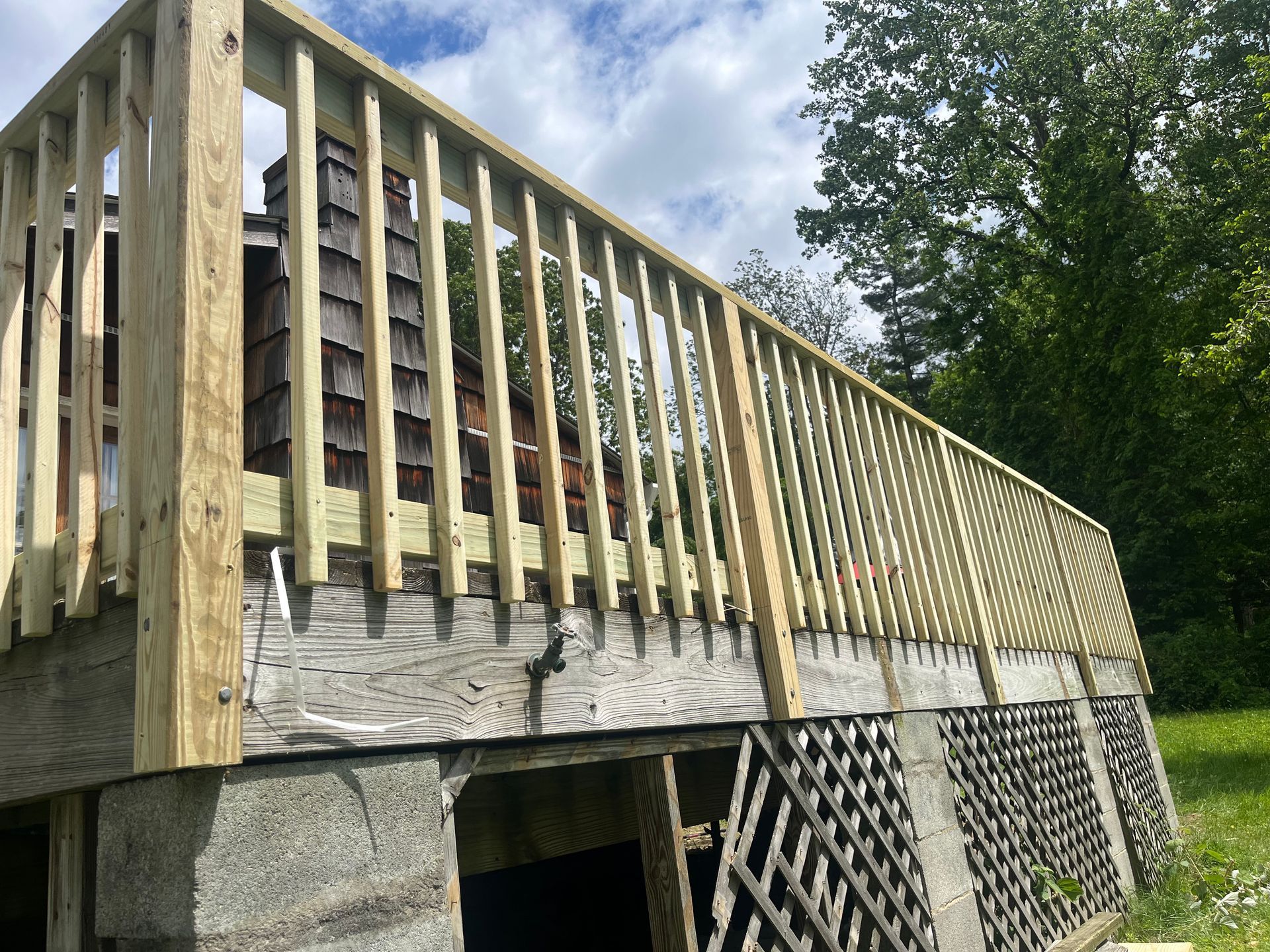 Wooden deck with railing attached to a concrete foundation, with a grassy yard and trees in the background.