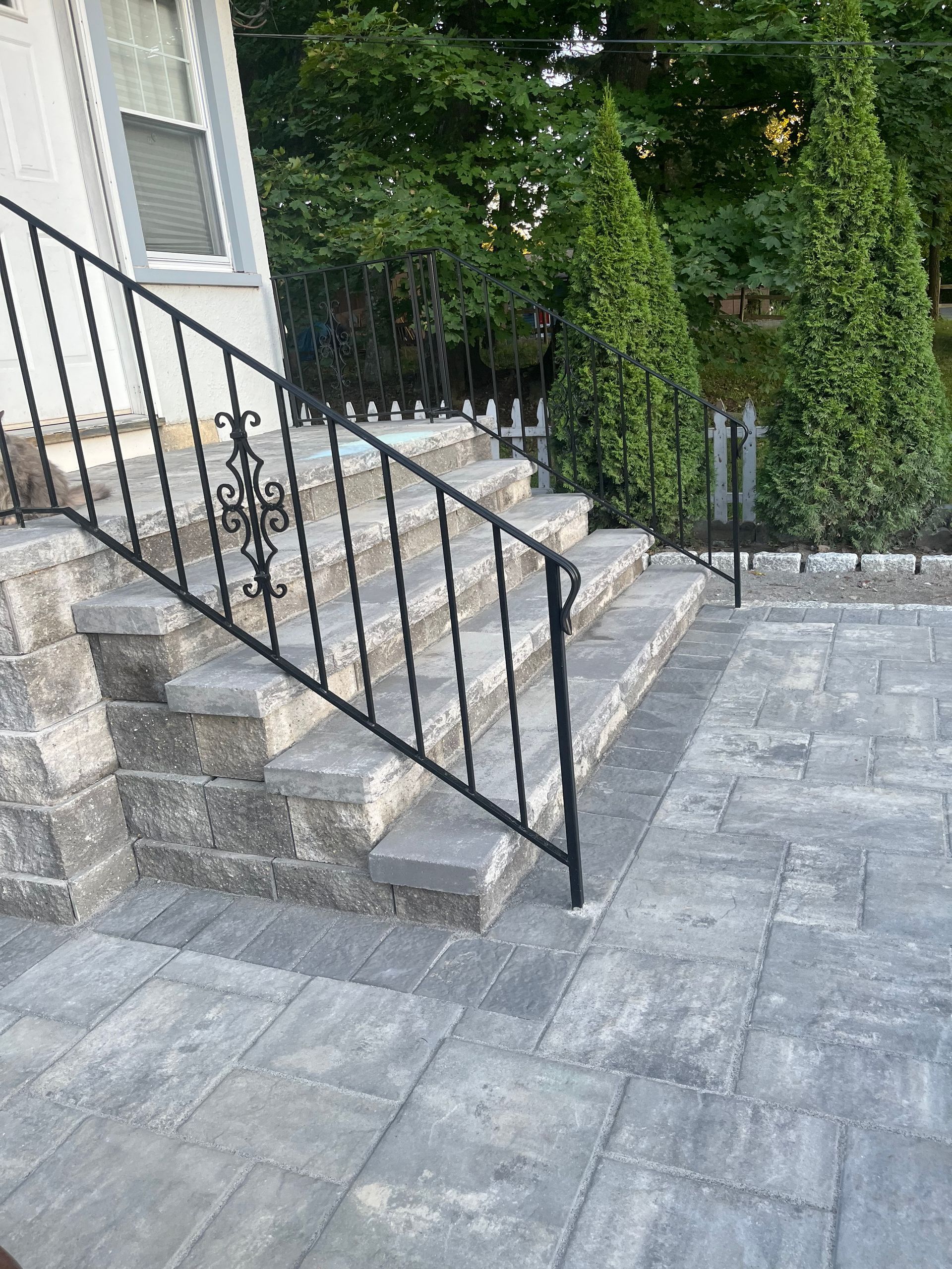 Stone steps and patio with black iron railing, leading to a white house with a green backdrop.