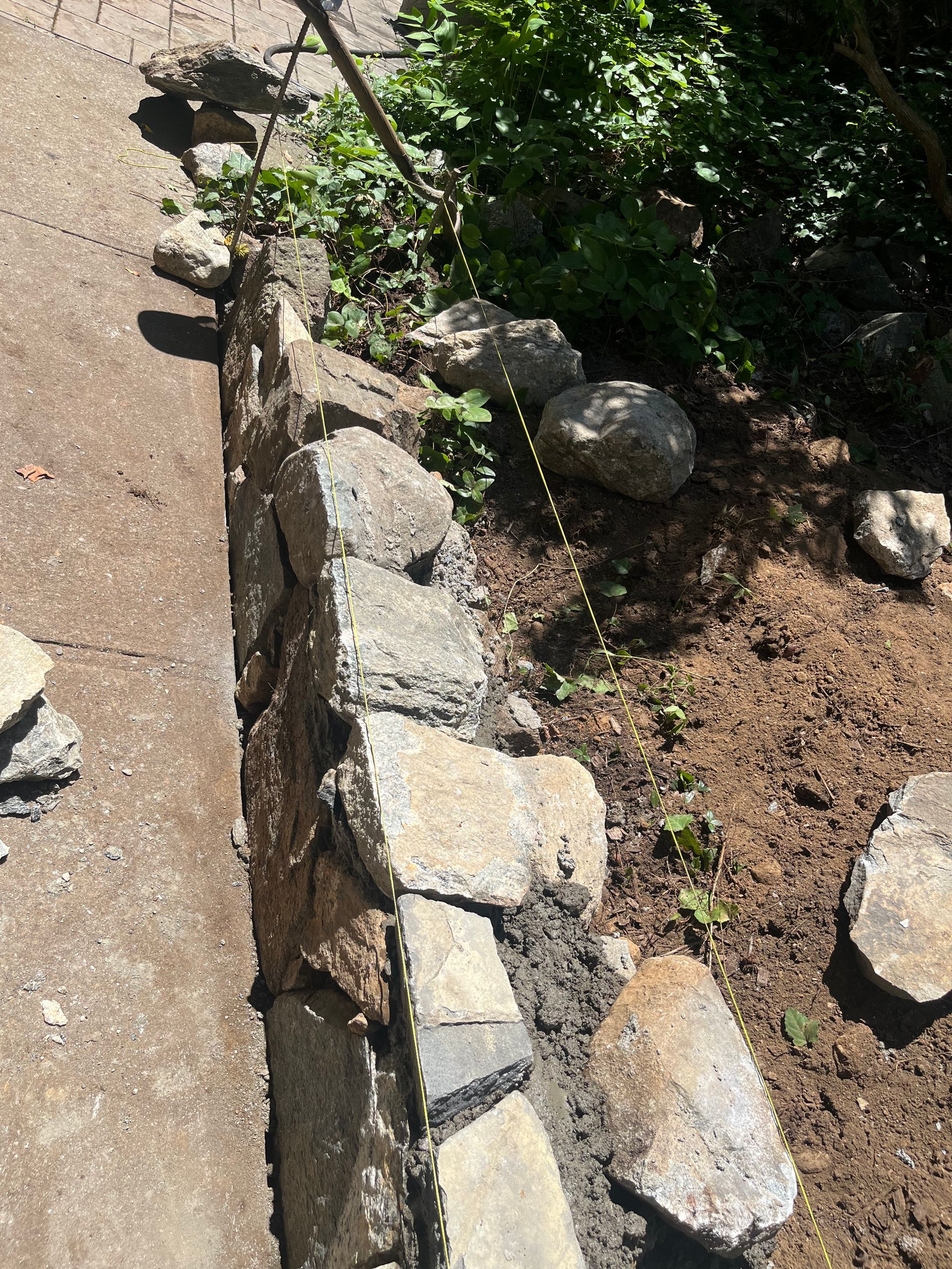A partially constructed stone retaining wall next to a dirt pathway. String lines guide the stones, surrounded by greenery and sunlight. Quiet and focused.