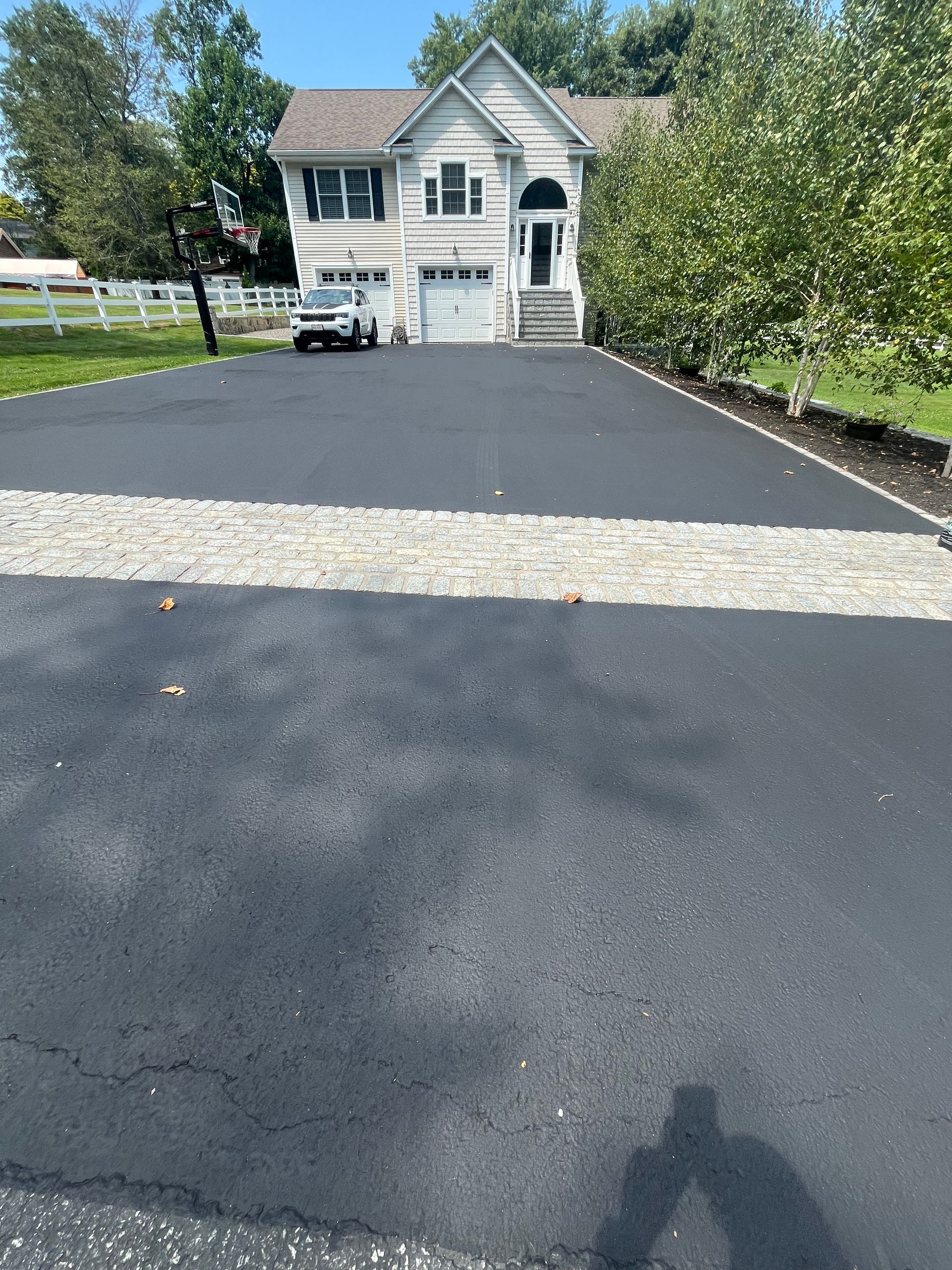 Black asphalt driveway leading to a two-story house with a light-colored stone insert.
