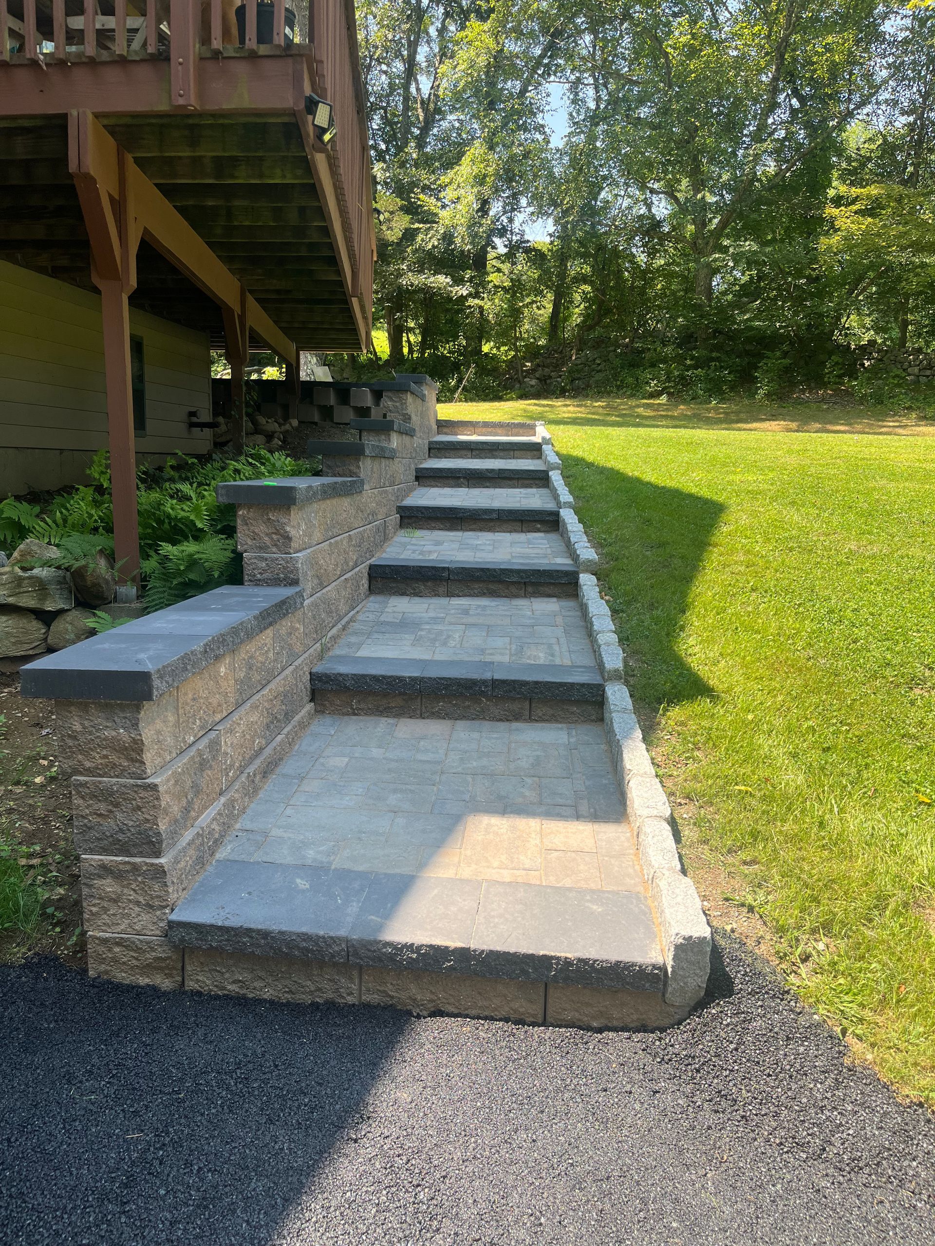 Stone stairs ascend alongside a wooden deck in a lush, sunlit garden. Trees and grass create a tranquil, serene atmosphere.