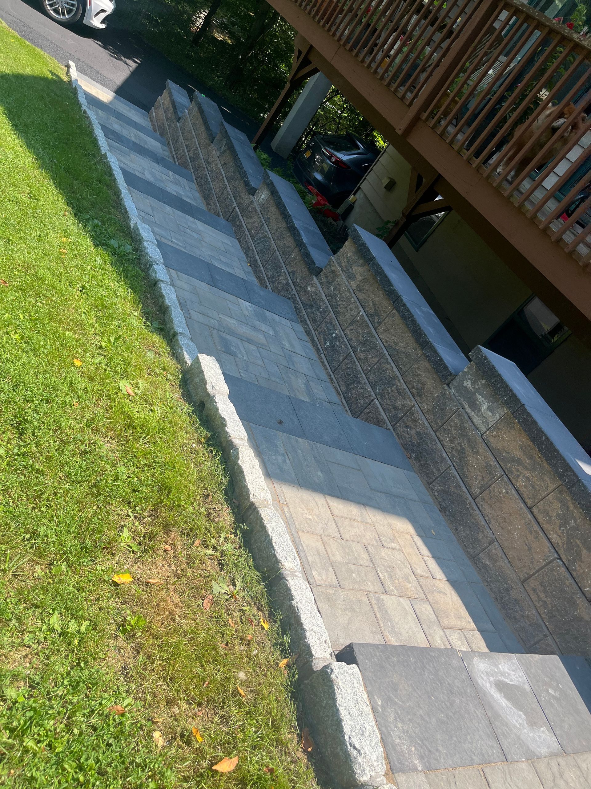 Outdoor stone staircase with retaining walls and paver steps, leading from a grassy yard to a driveway beneath a wooden deck.