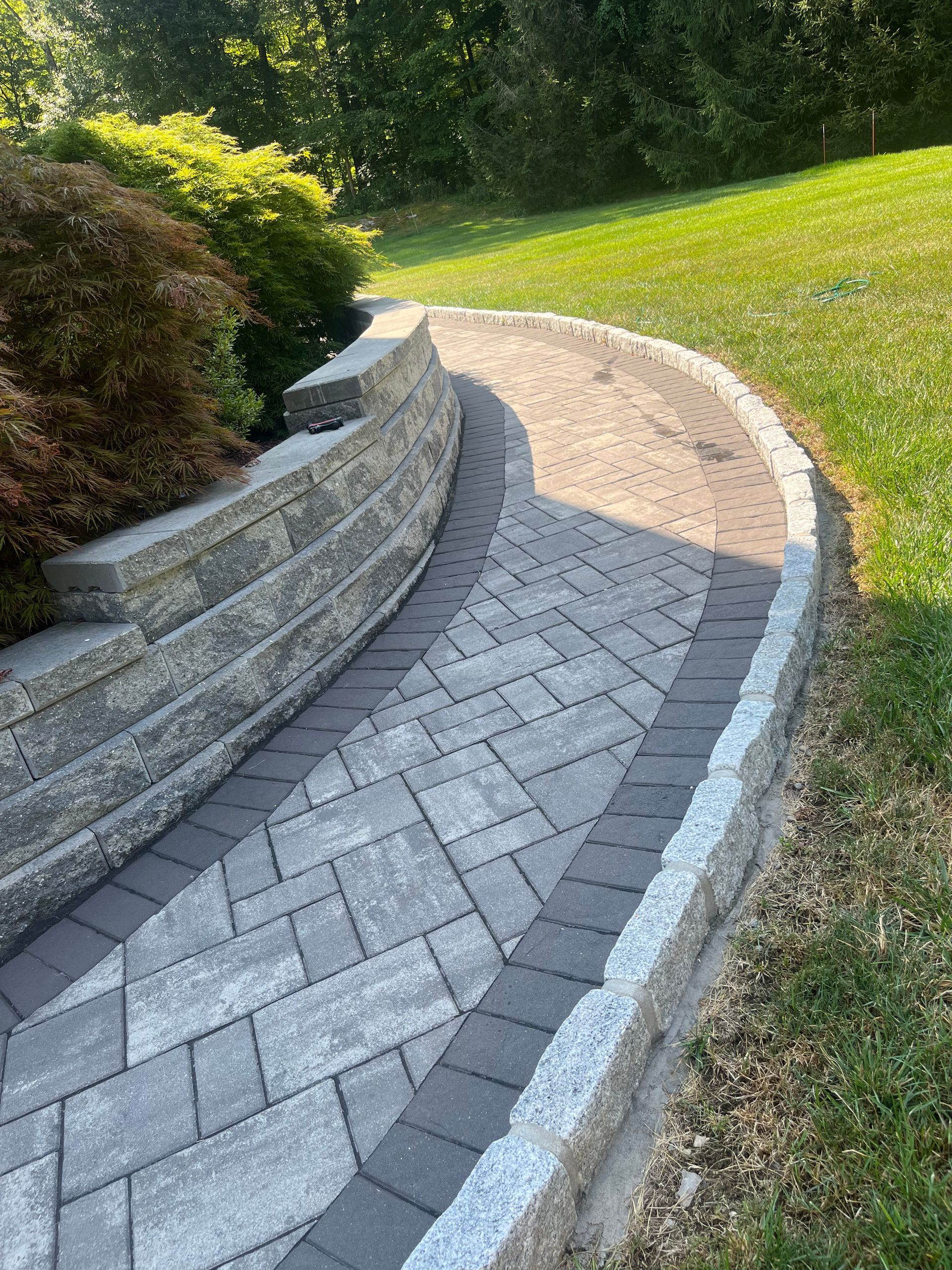 Curved stone walkway with interlocking pavers and a tiered retaining wall, bordered by grass and landscaped shrubs.