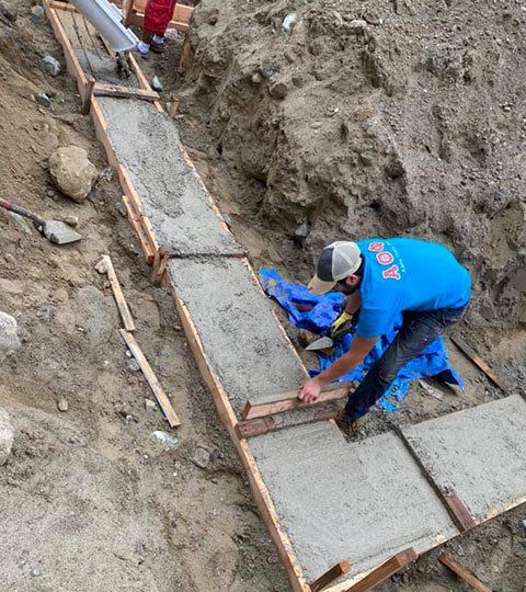 Man pouring concrete into wooden forms on a sloped terrain. Earth tones dominate the scene.