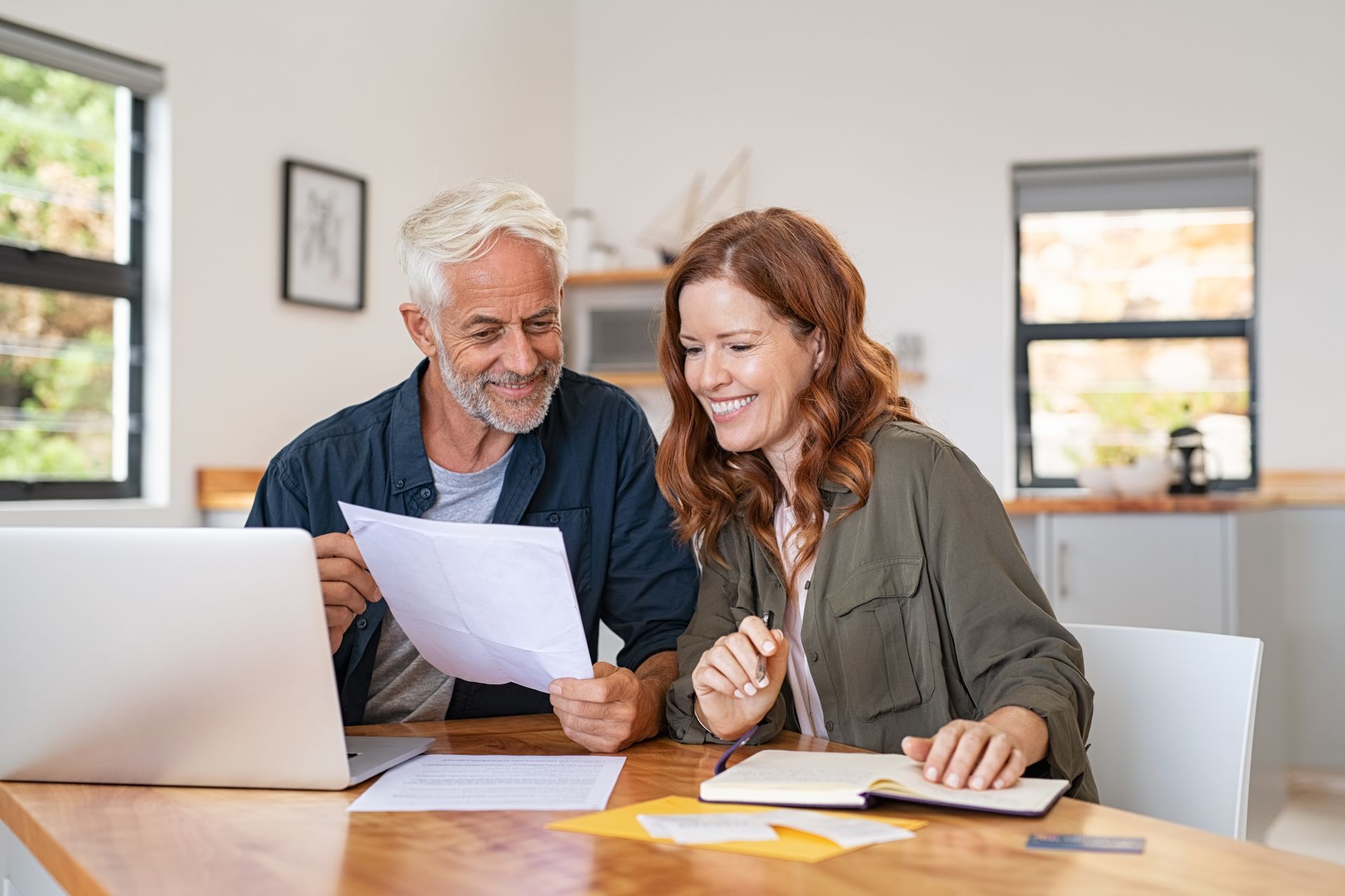 Couple reviewing paperwork together at a table with a laptop, smiling in a bright room.