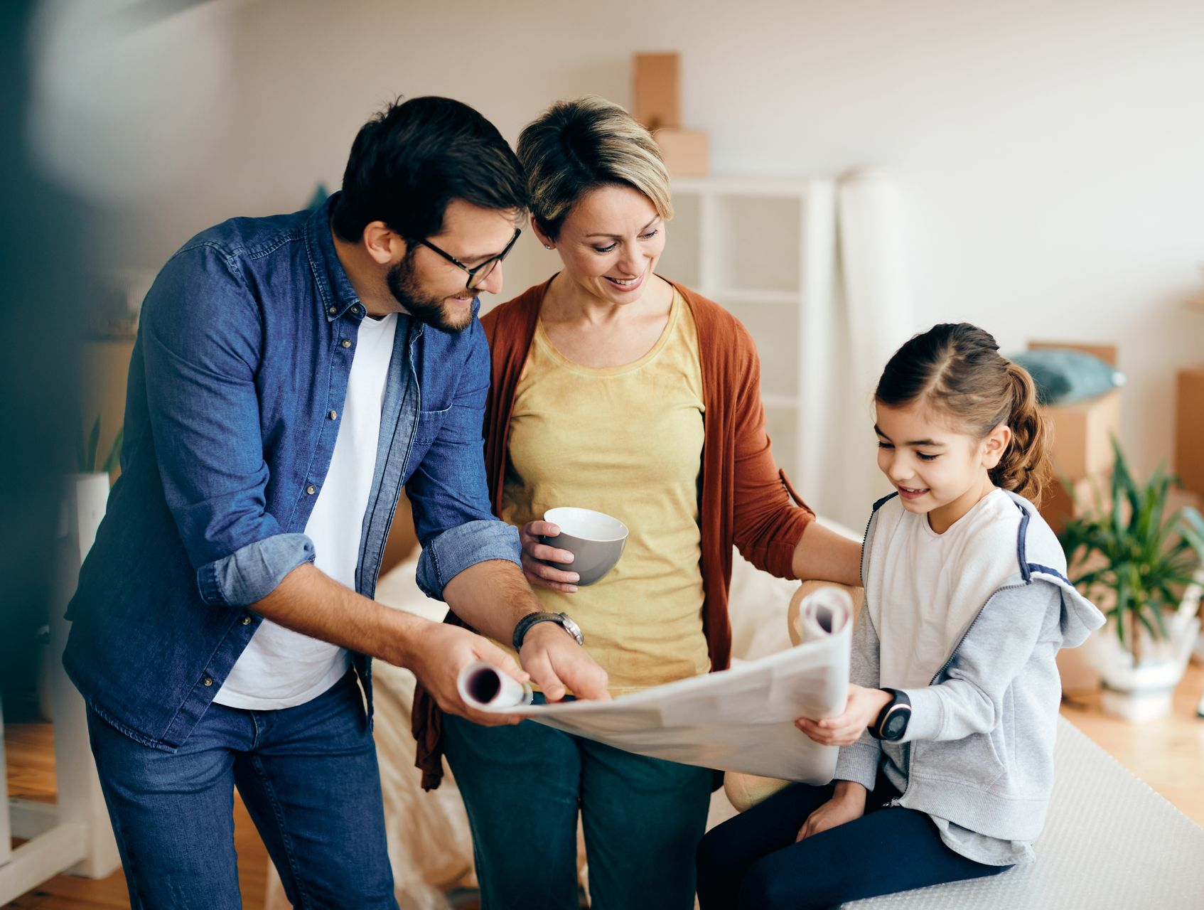 Family, looking at a rolled-up paper, smiles in a room. A man points, a woman holds a cup, and a girl sits.