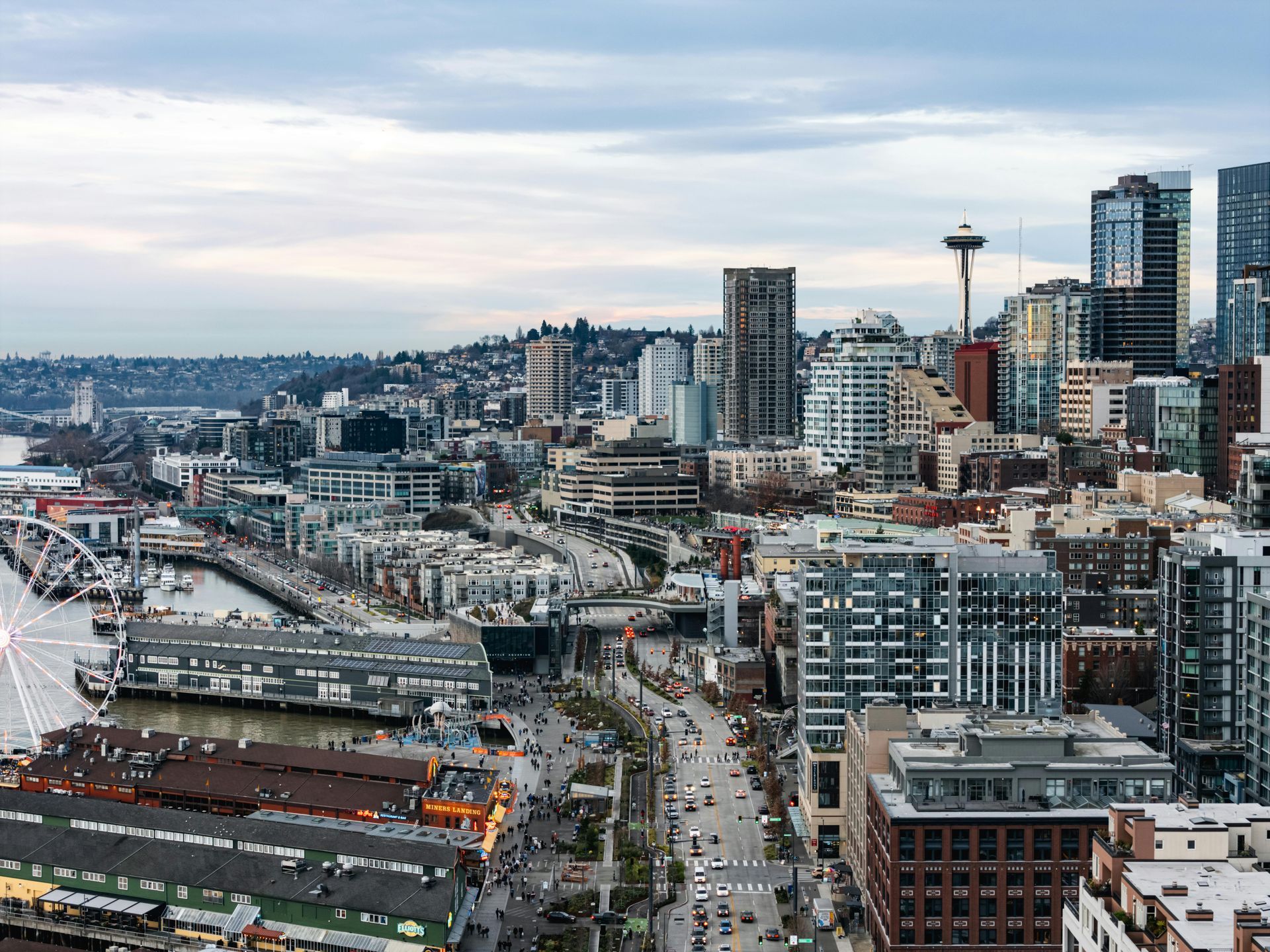 Seattle cityscape with waterfront, buildings, and Space Needle under a cloudy sky.
