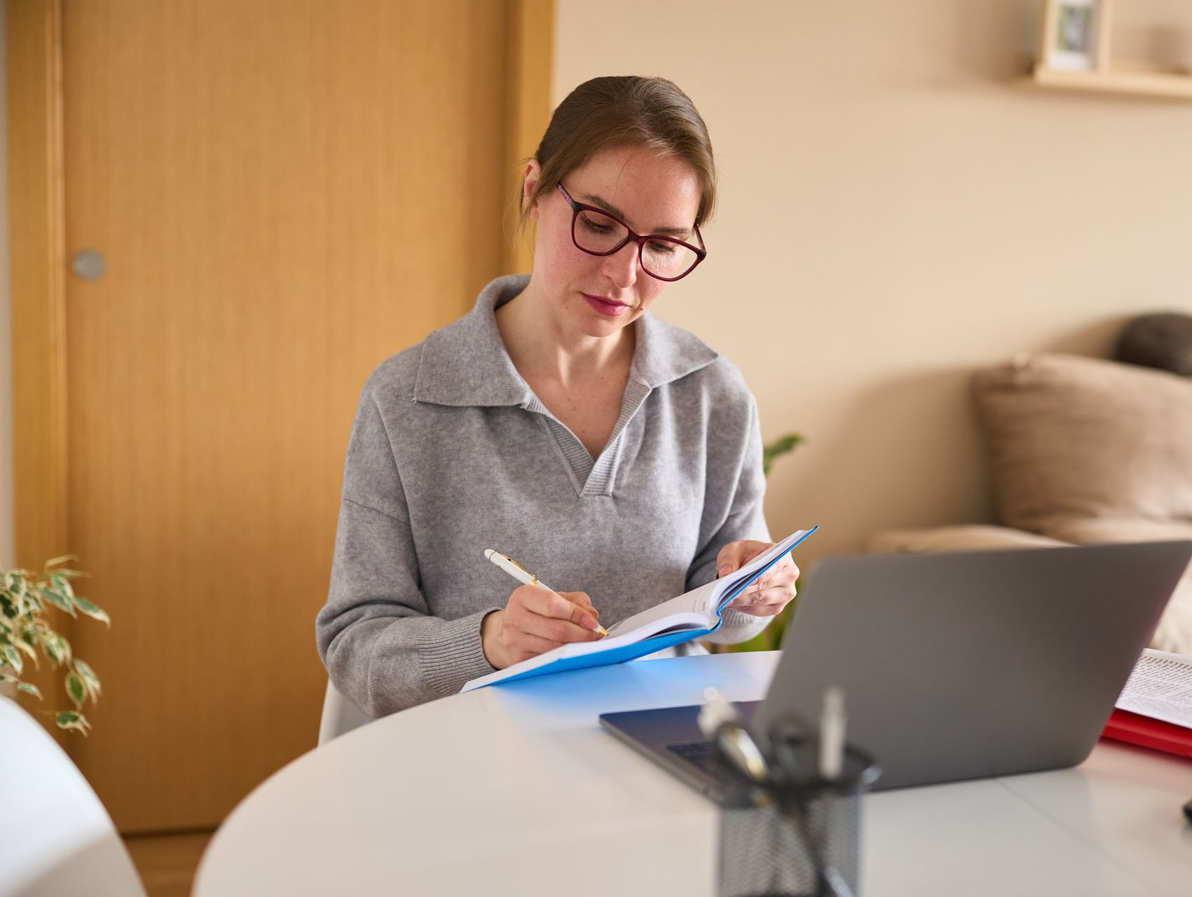 Woman in glasses writing in notebook at a table with laptop, pen, and desk supplies.