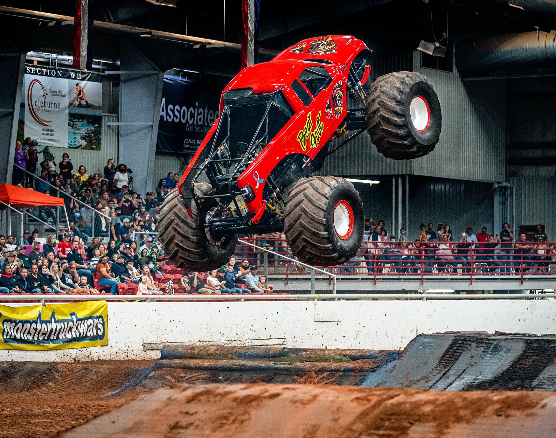 Red monster truck in mid-air above a dirt track, with a crowd watching.