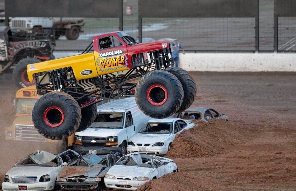Monster truck, Carolina Crusher, jumps over cars at a dirt track event. Yellow, red, and blue paint scheme.