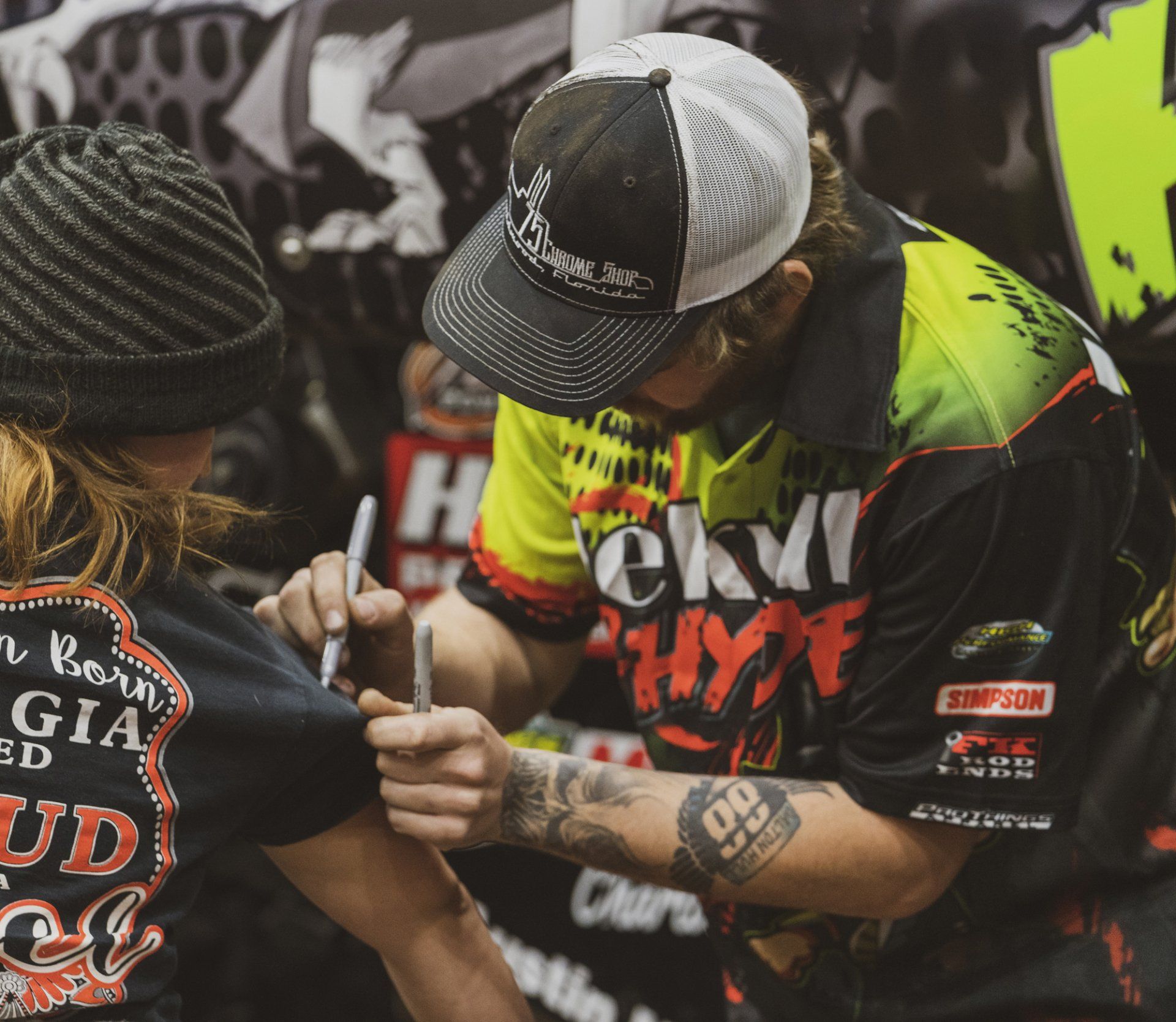 Man in racing gear signing a shirt, indoors at an event. He wears a trucker hat, black, yellow, and red shirt.