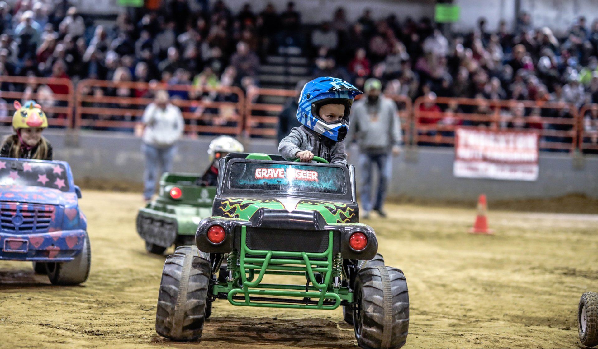 Children driving toy monster trucks in an arena, wearing helmets.