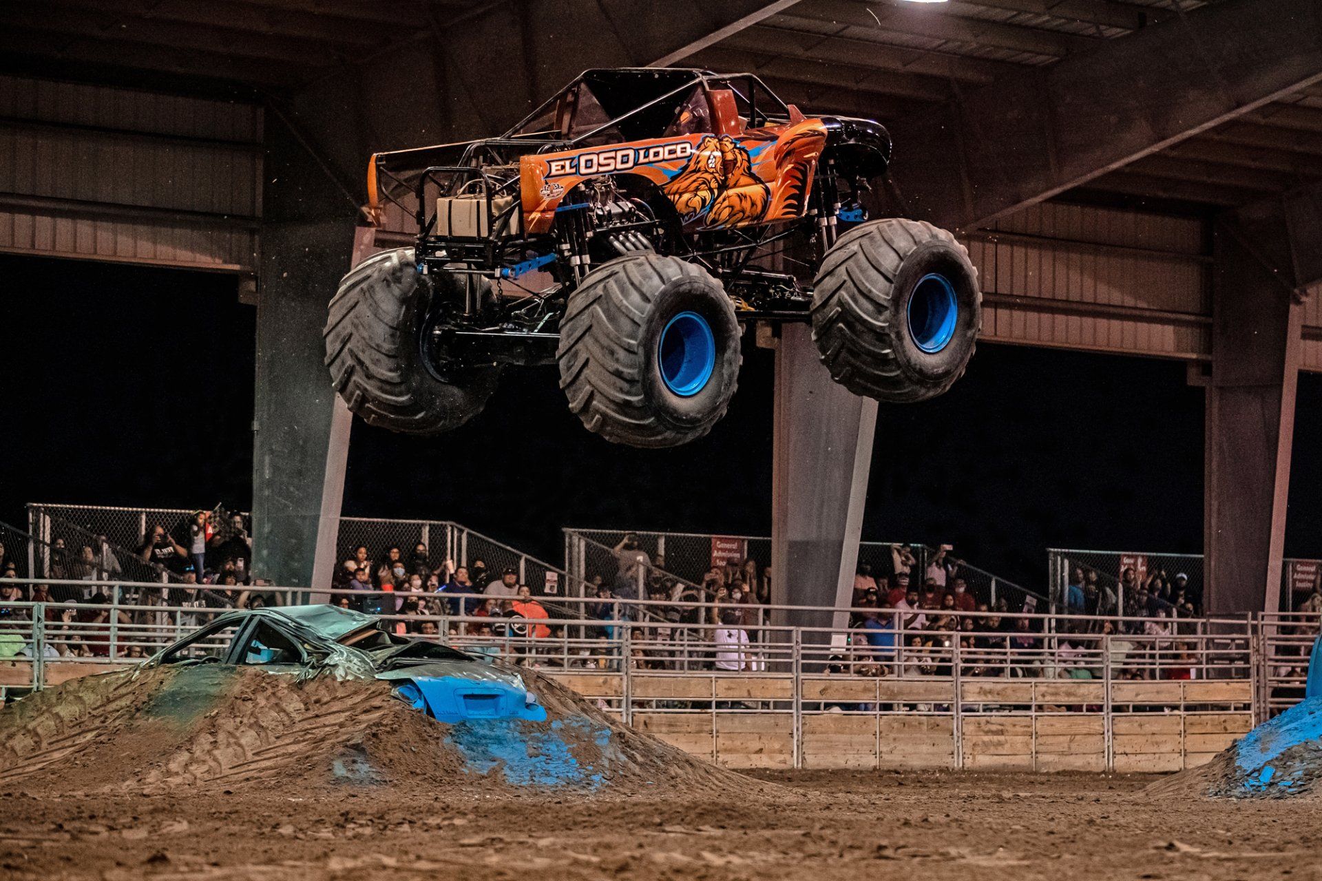 Monster truck in mid-air above a dirt ramp and crushed car at an arena event.